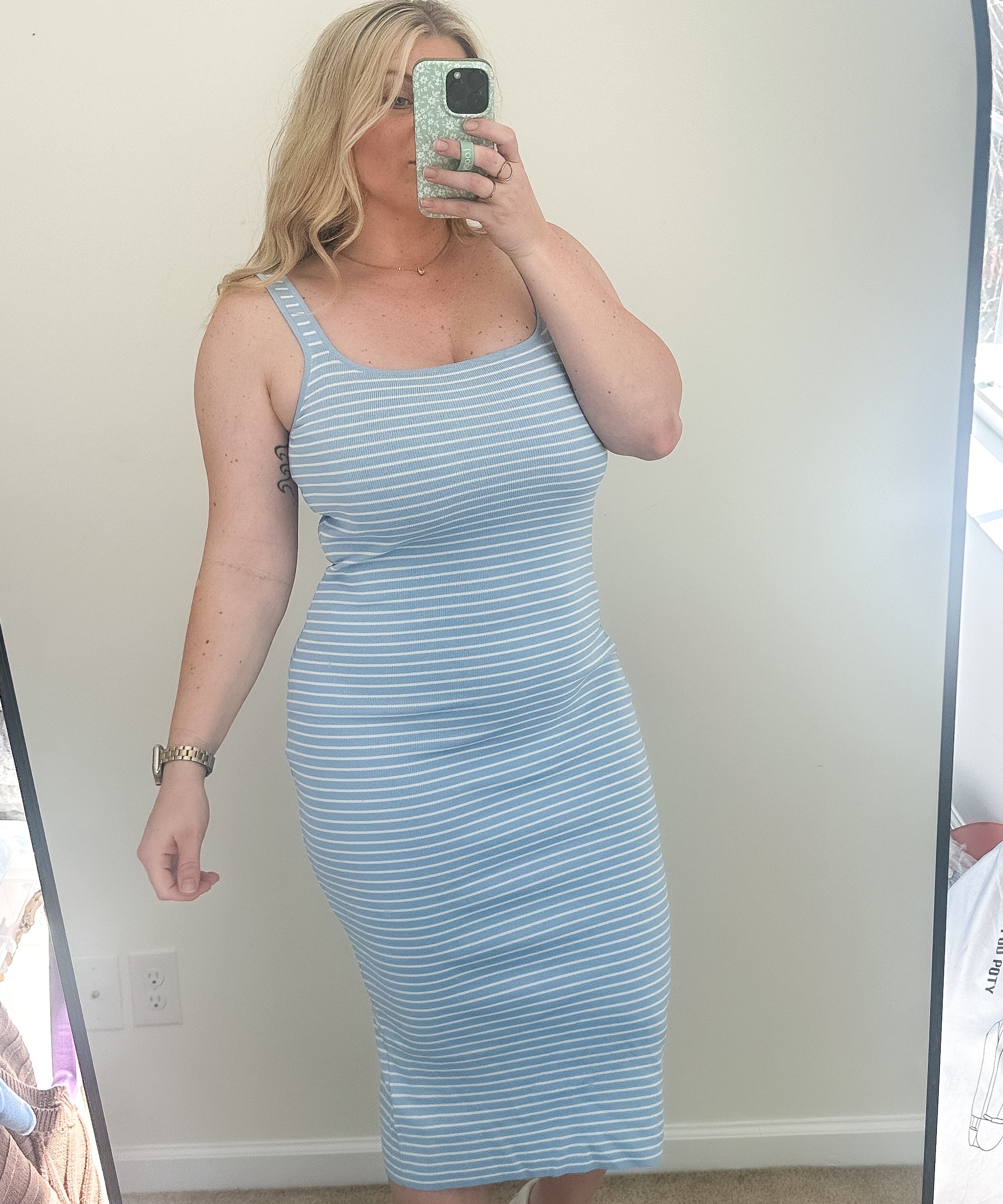 Woman taking a mirror selfie wearing a light blue striped dress in a room with a window.