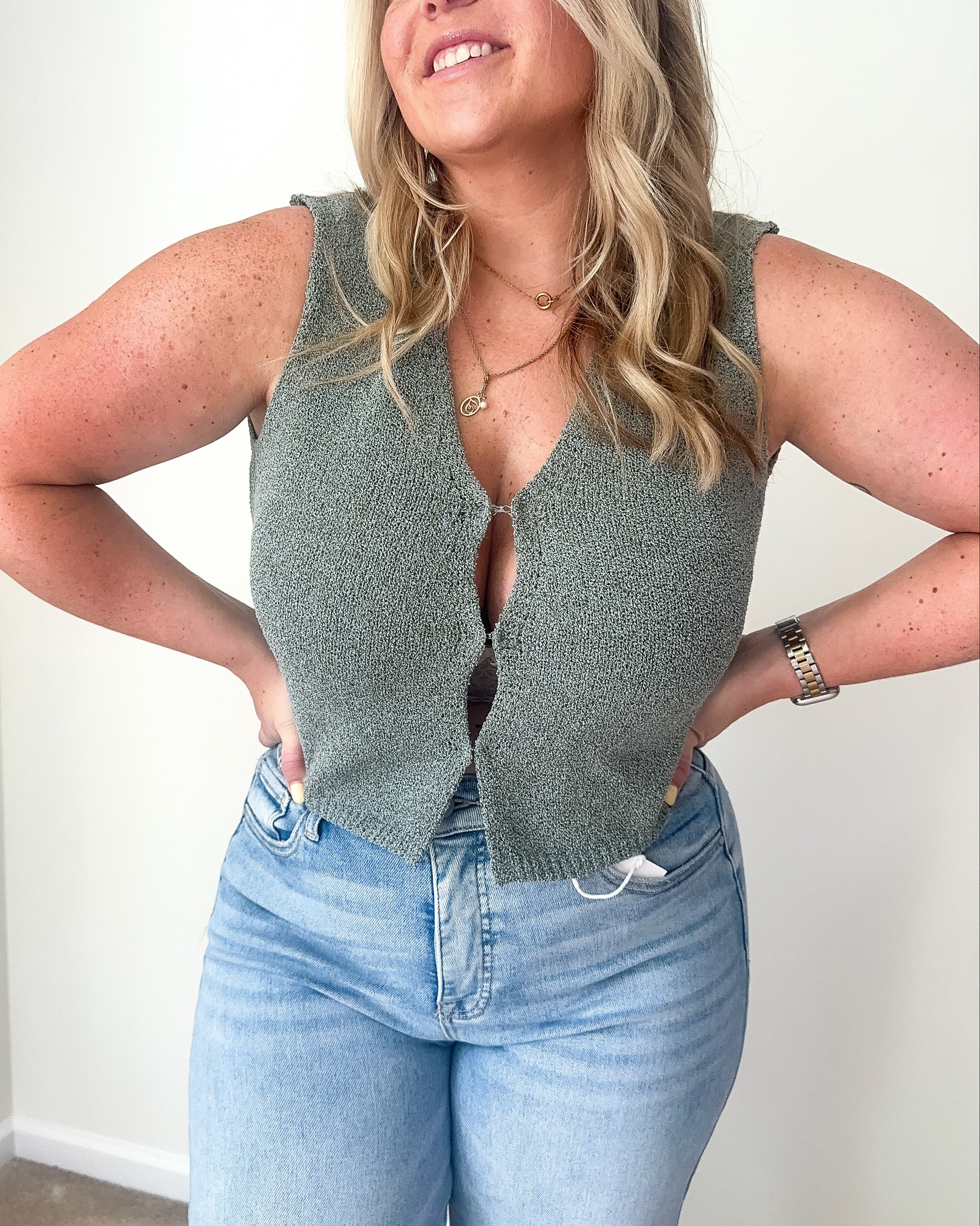 A sleeveless olive green knit vest cardigan with a deep V-neckline and hook-and-eye closure.