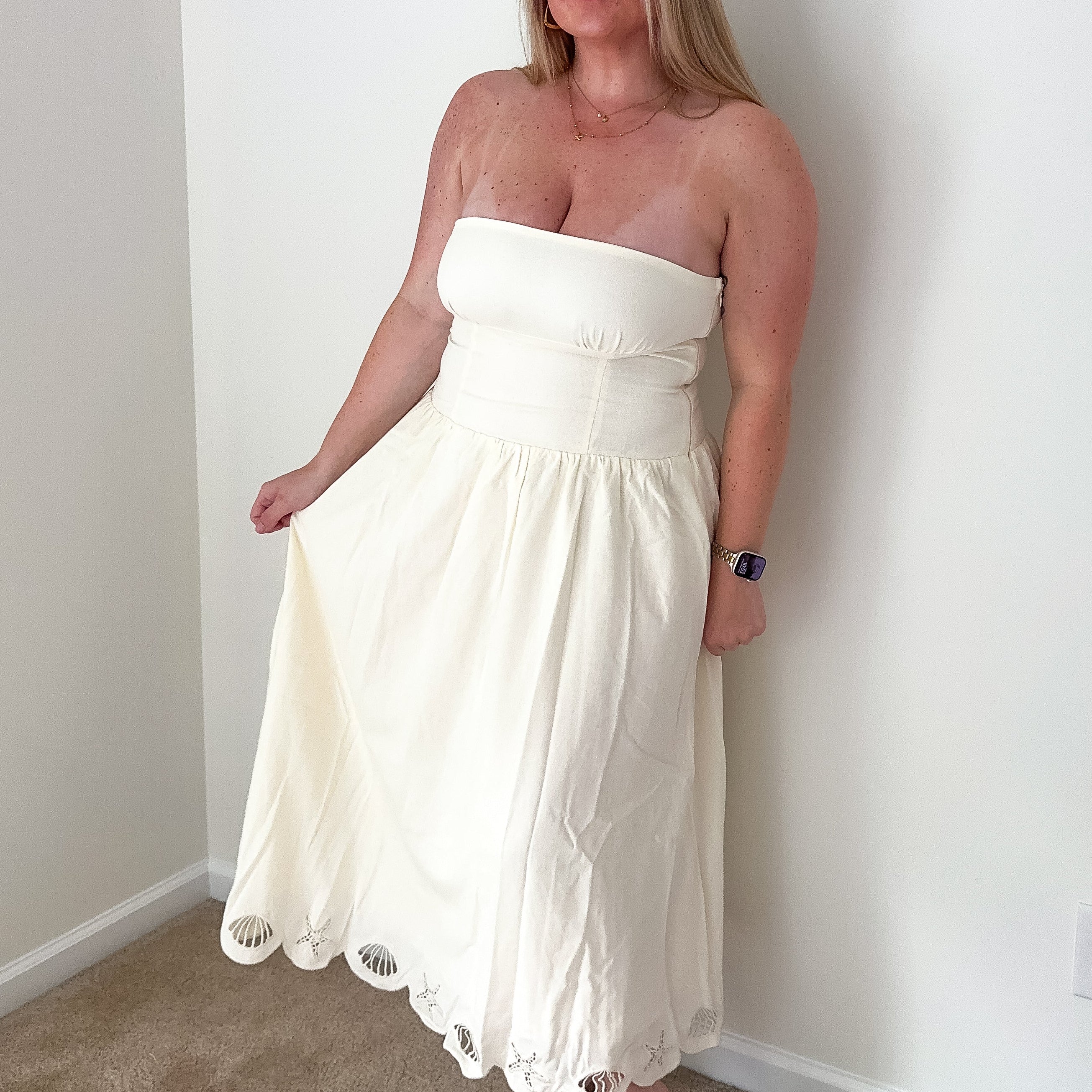 Woman wearing a white strapless dress standing against a plain wall.