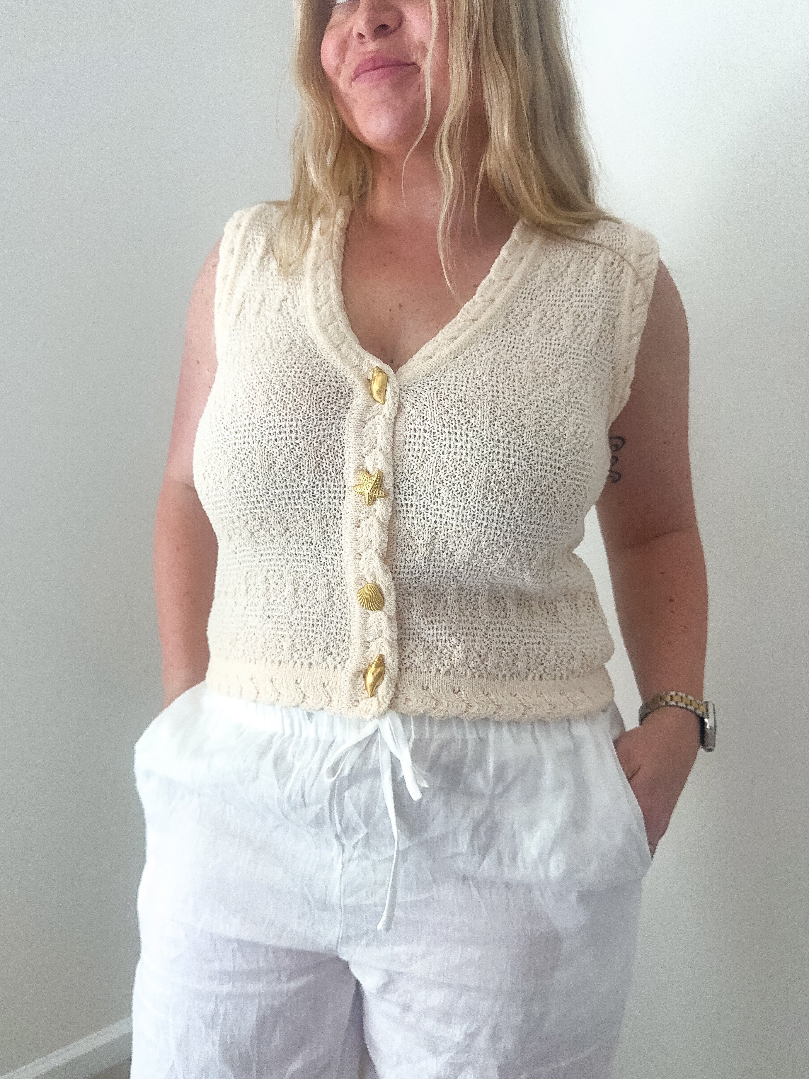 Woman wearing a cream knitted vest with buttons against a plain background