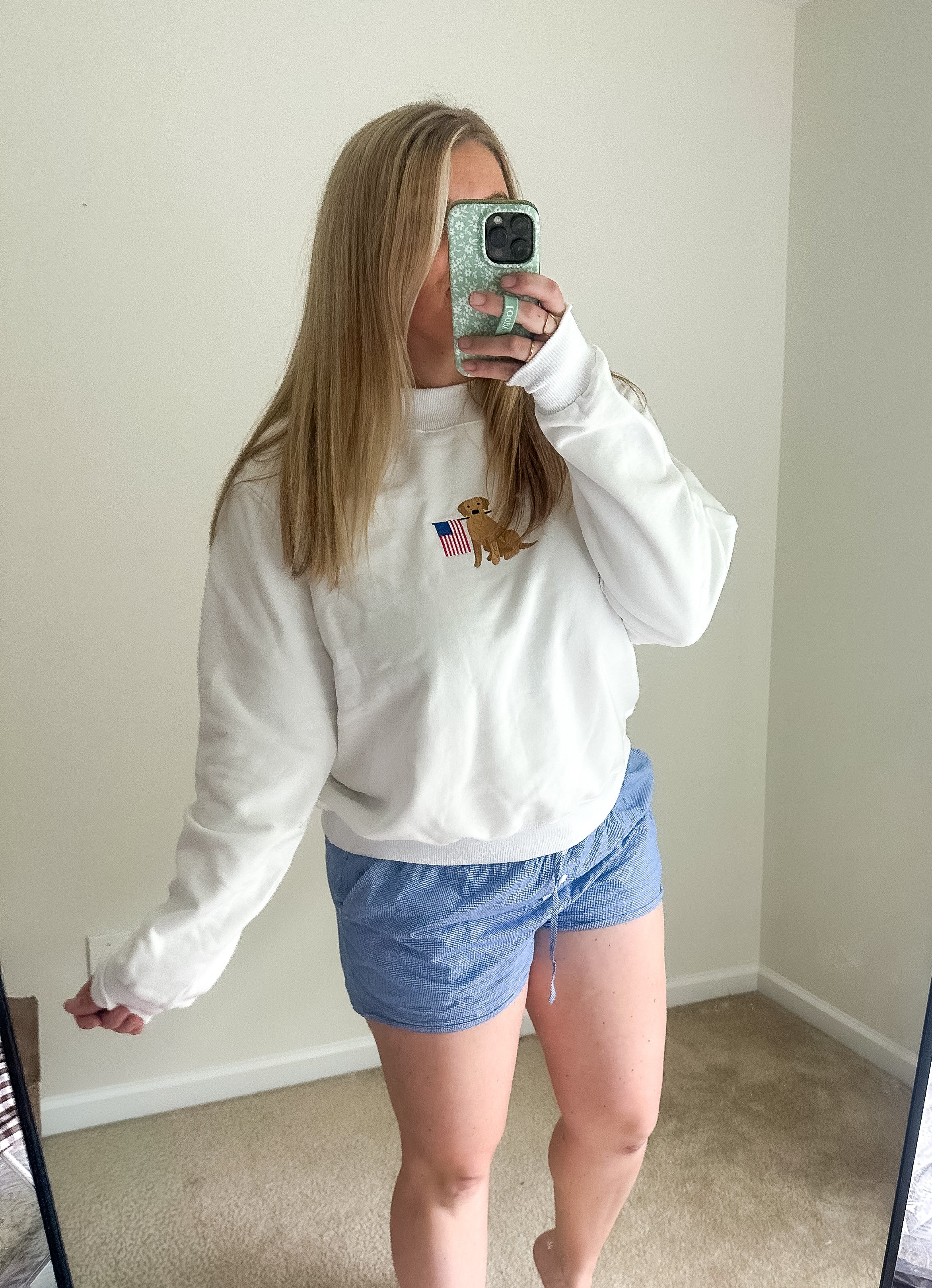 Person wearing a white sweatshirt with a logo and blue shorts taking a mirror selfie.