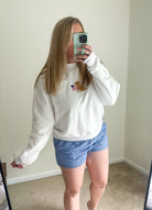 Person wearing a white sweatshirt with a logo and blue shorts taking a mirror selfie.