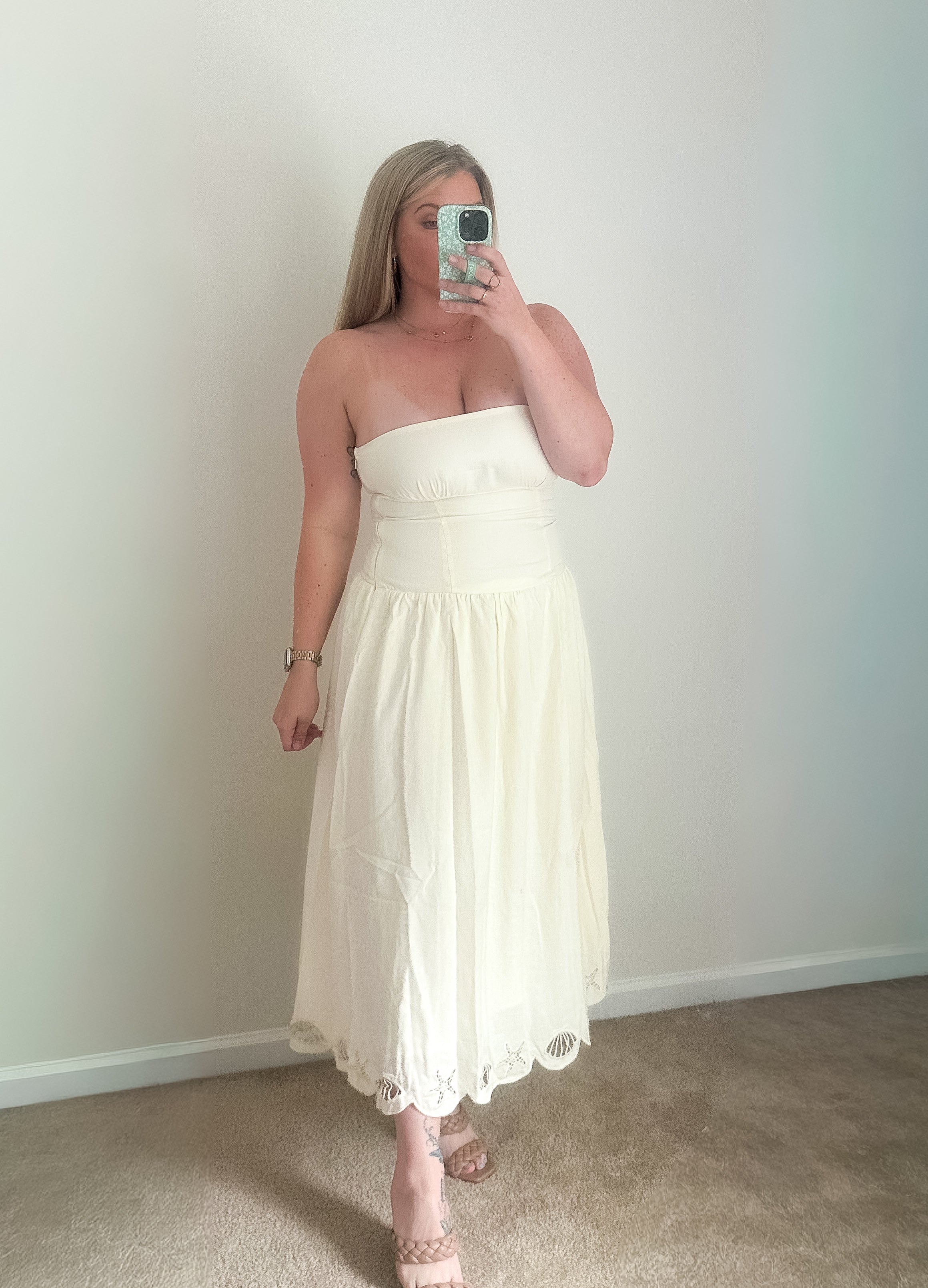 Woman wearing a white strapless dress taking a mirror selfie against a plain wall.