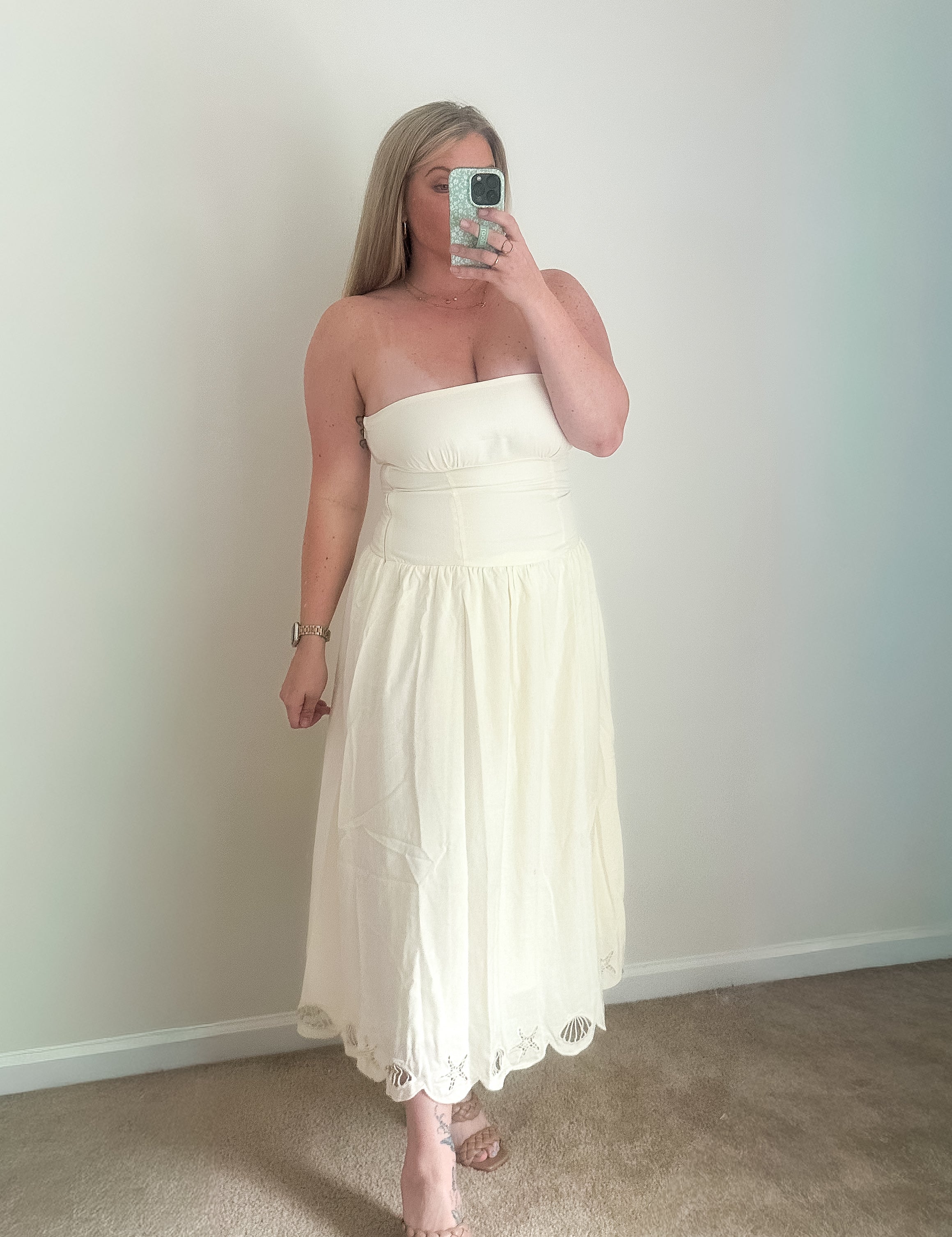 Woman wearing a white strapless dress taking a mirror selfie against a plain wall.