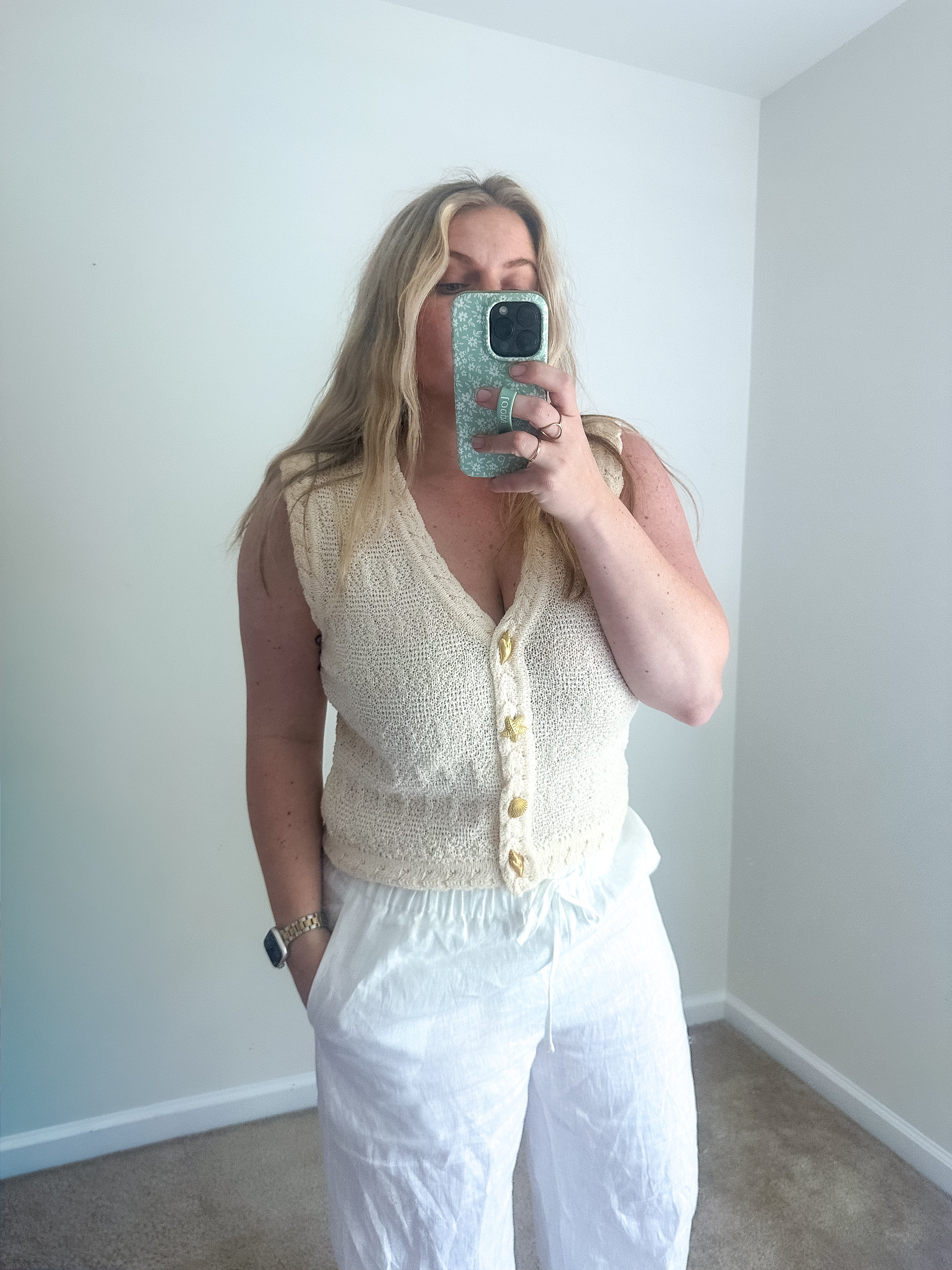 Woman taking a mirror selfie wearing a white sleeveless top and pants in a room with white walls.