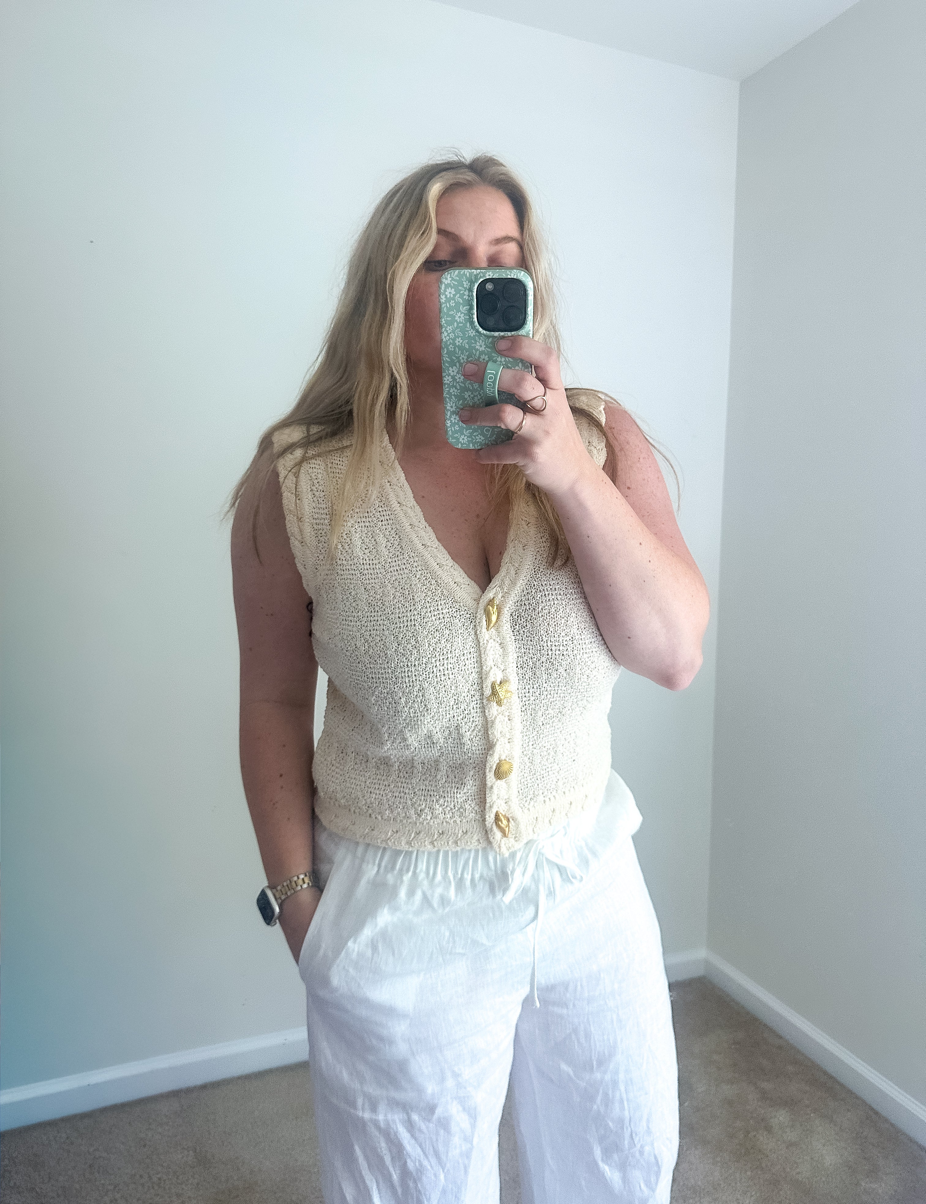 Woman taking a mirror selfie wearing a white sleeveless top and pants in a room with white walls.