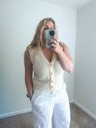 Woman taking a mirror selfie wearing a white sleeveless top and pants in a room with white walls.