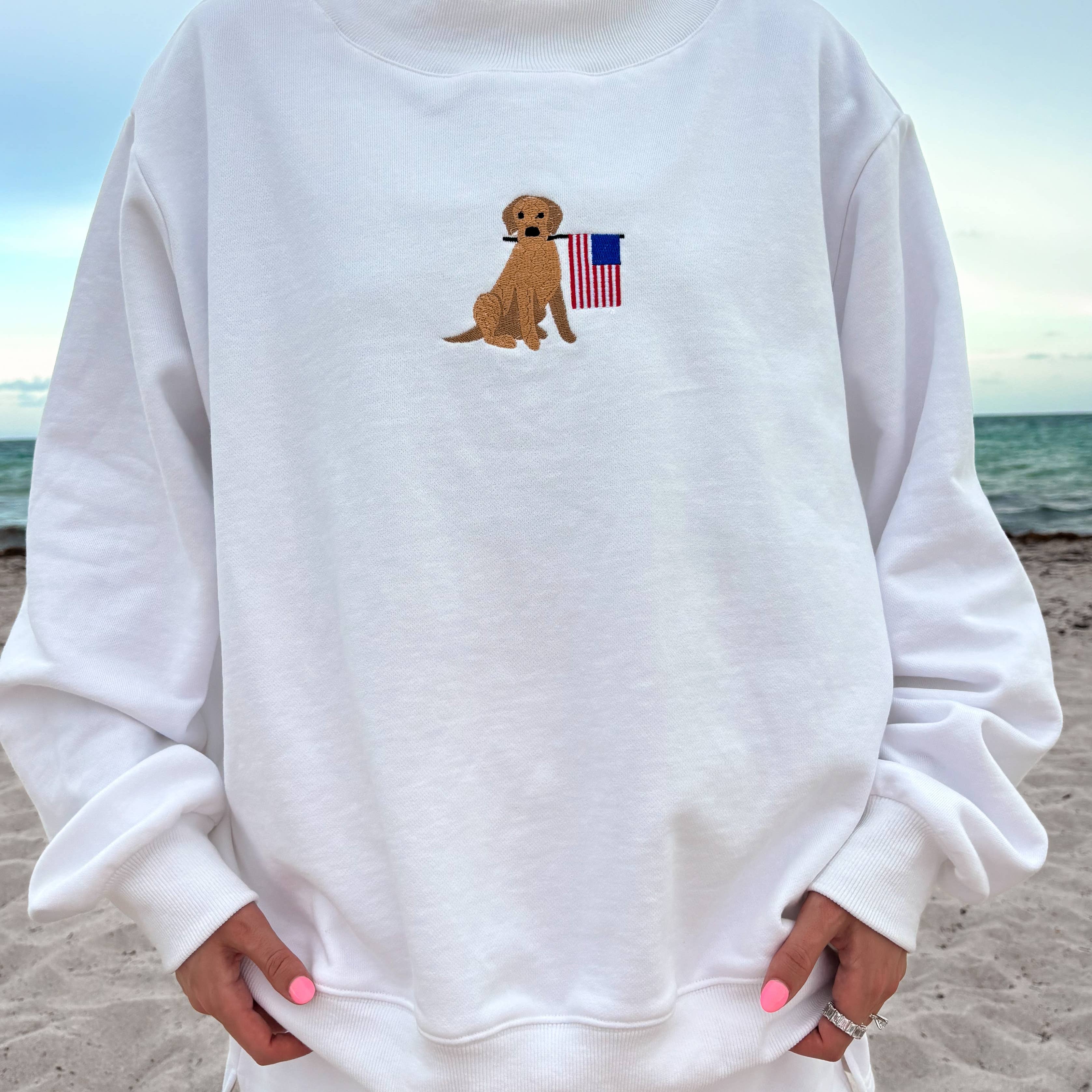 Person wearing a white sweatshirt with a dog and American flag design on a beach.