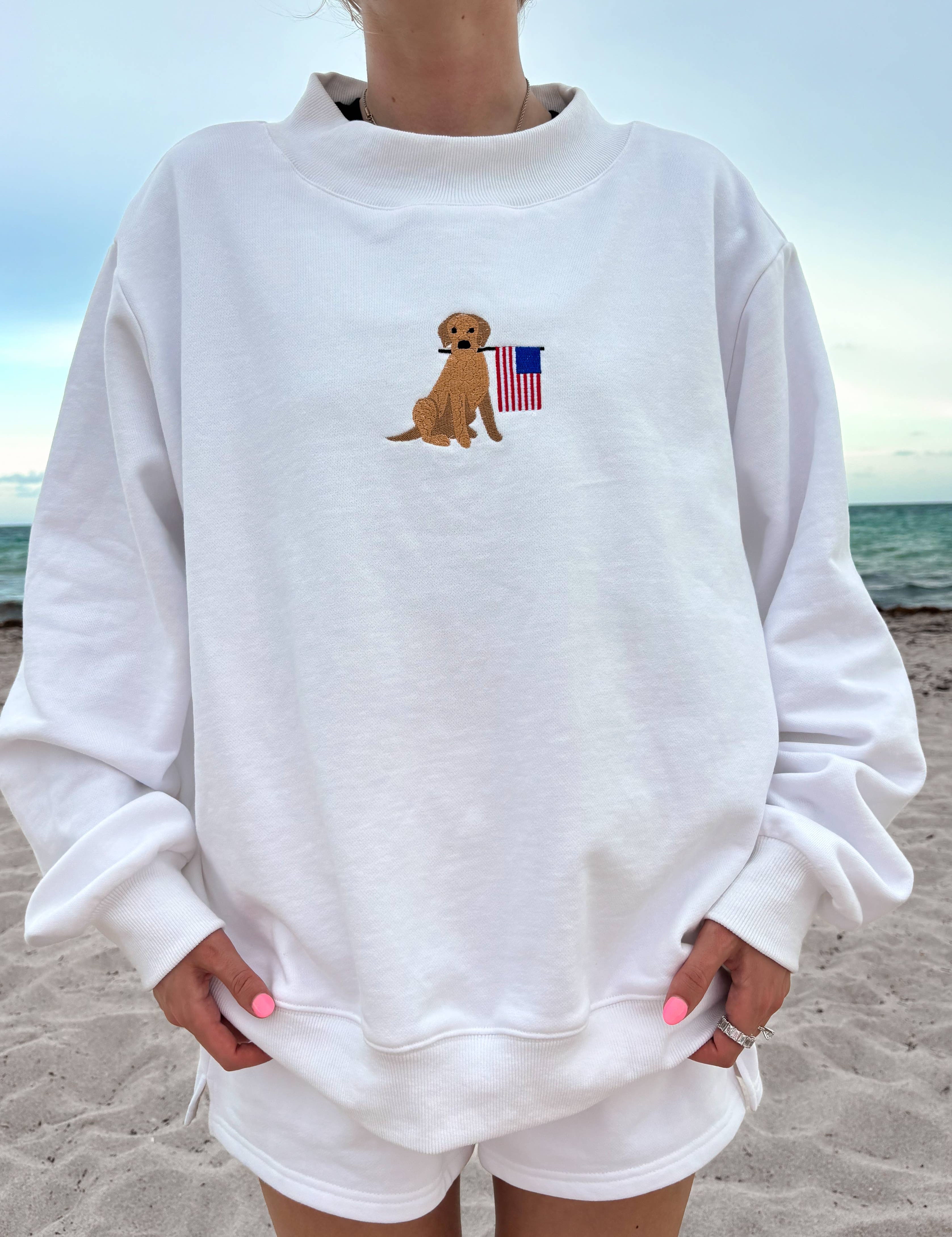 Person wearing a white sweatshirt with a dog and American flag design on a beach.