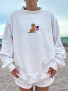 White mockneck sweatshirt with an embroidered golden pup holding a flag, worn on the beach.