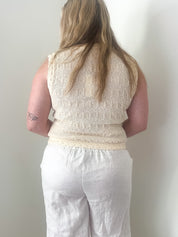 Person wearing a beige sleeveless top and white pants against a plain wall.
