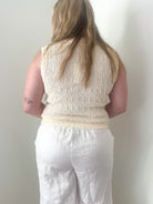 Person wearing a beige sleeveless top and white pants against a plain wall.