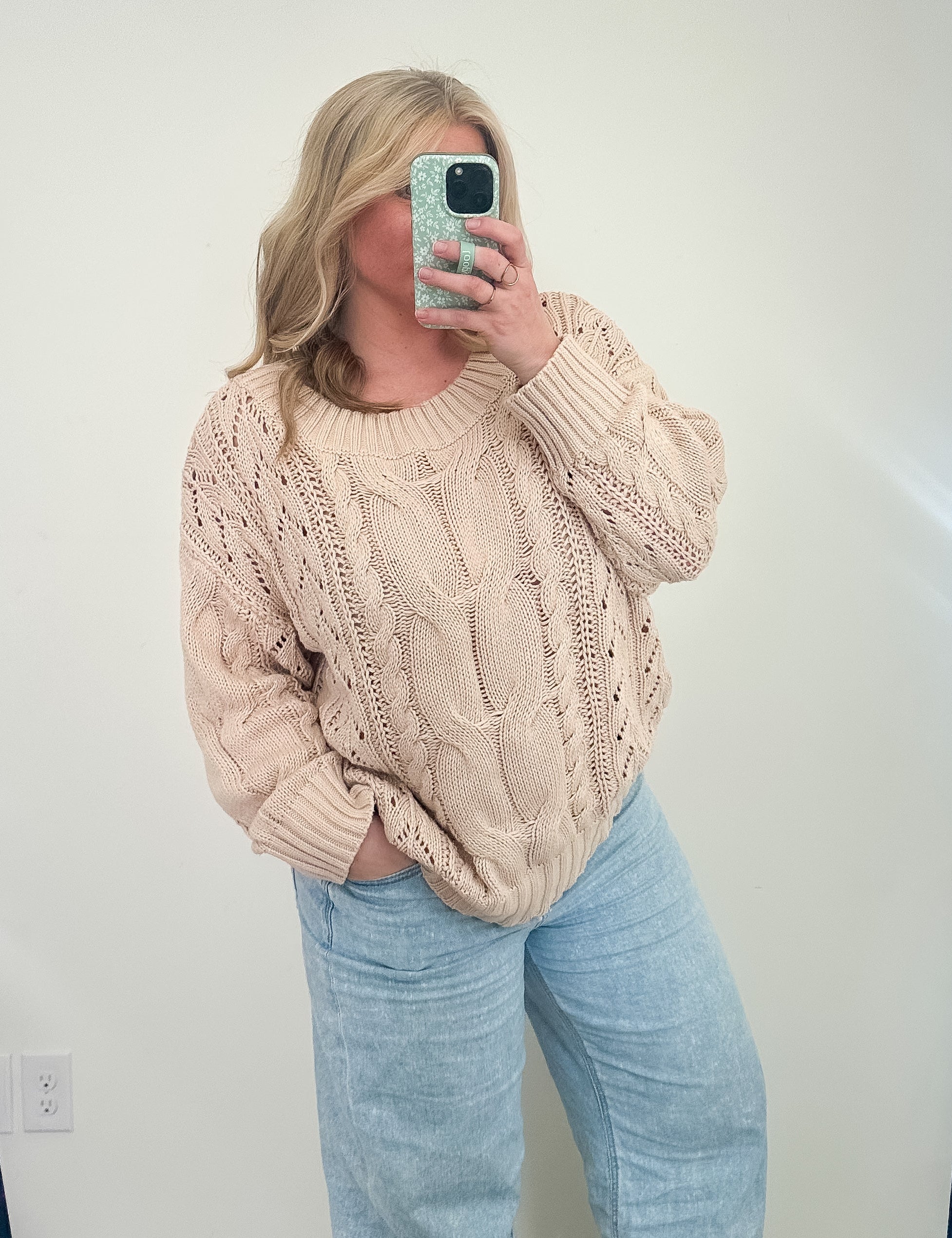 Person wearing a beige cable knit sweater and light blue jeans taking a mirror selfie.