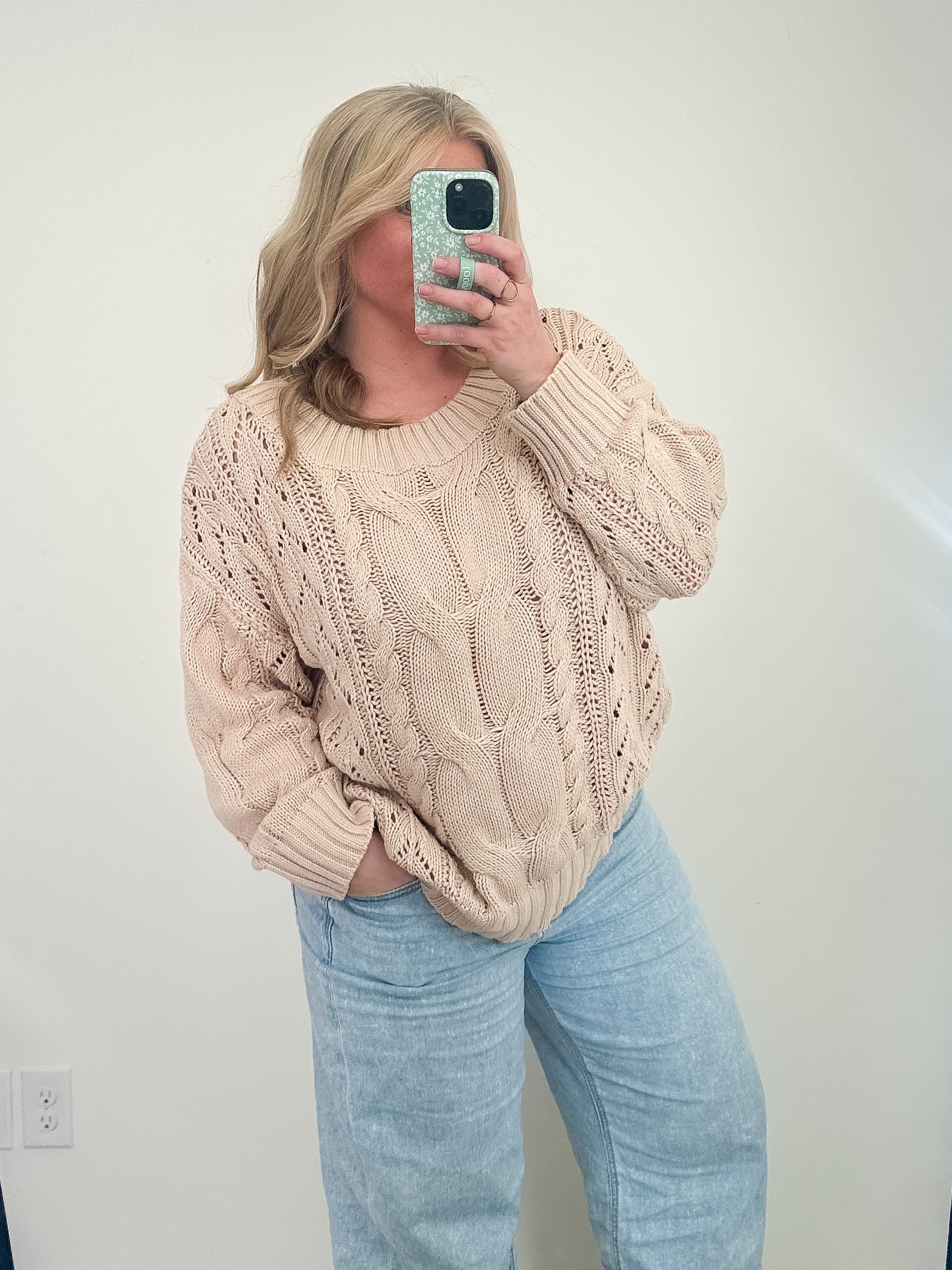 Person wearing a beige cable knit sweater and light blue jeans taking a mirror selfie.