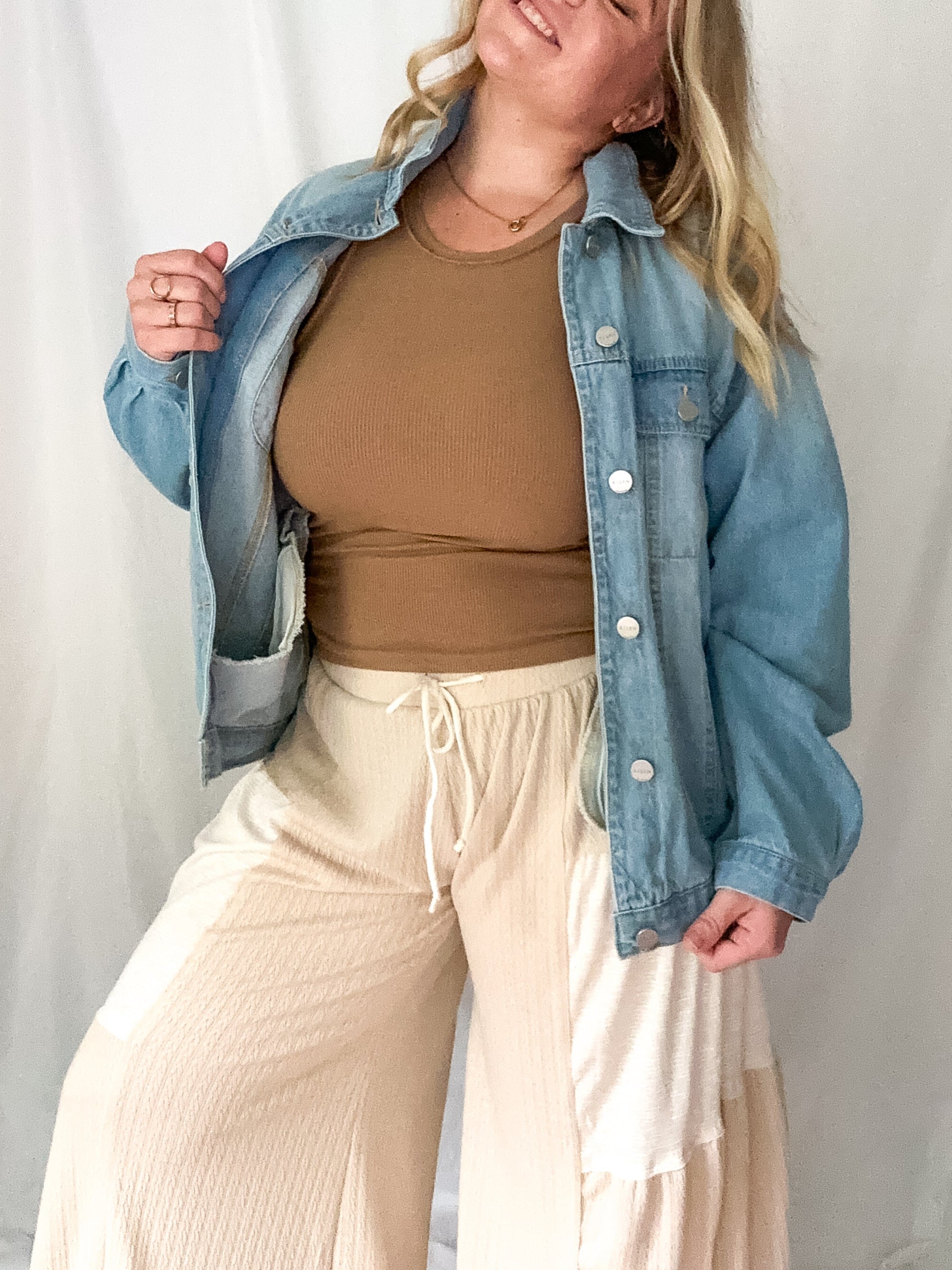 Woman wearing a denim jacket, brown top, and beige pants against a white background