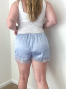 Person wearing a white tank top and light blue shorts with lace trim against a plain wall.