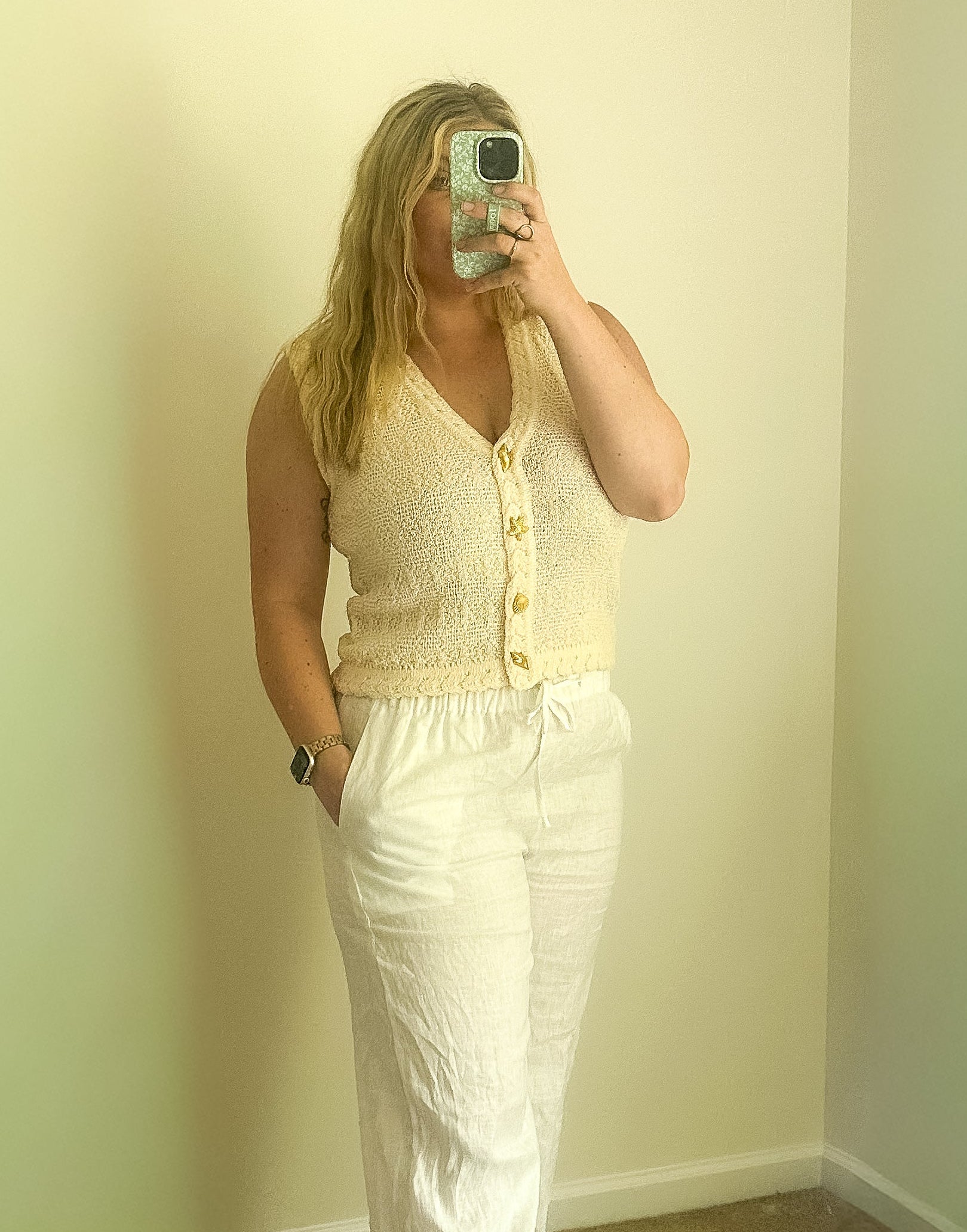 Person taking a mirror selfie wearing a yellow sleeveless top and white pants in a room with light green walls.