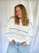 A white crochet sweater with blue, teal, and green stripes, featuring a pointelle design and relaxed fit.