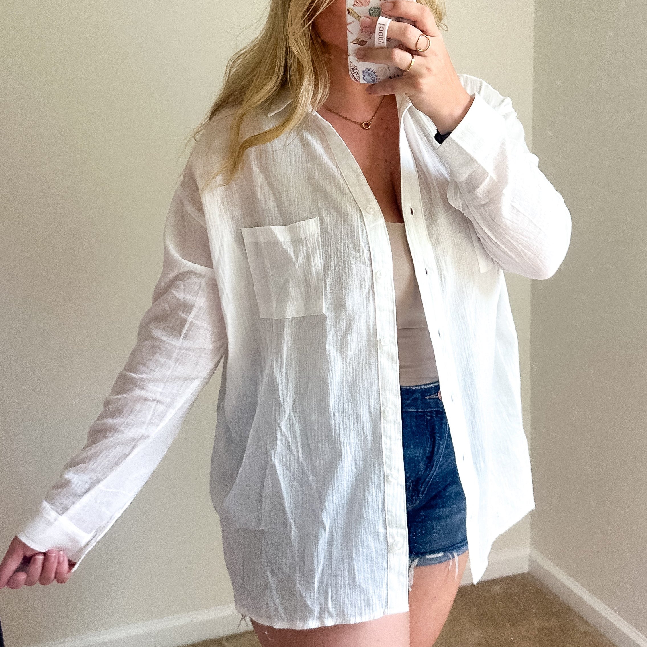 A woman takes a mirror selfie wearing an oversized white long-sleeve button-up shirt over a white top and denim shorts in a room with beige walls and carpet.