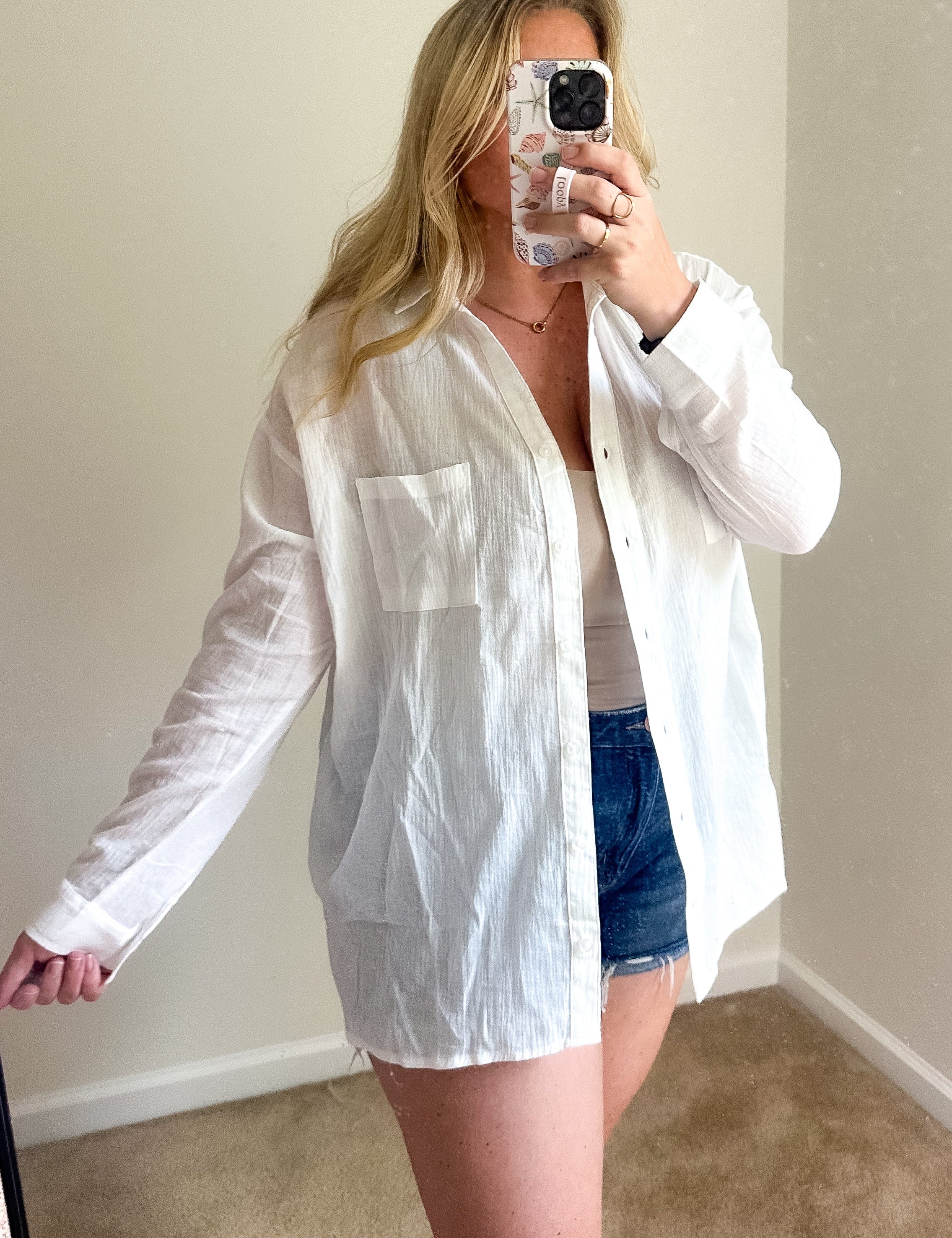 A woman takes a mirror selfie wearing an oversized white long-sleeve button-up shirt over a white top and denim shorts in a room with beige walls and carpet.