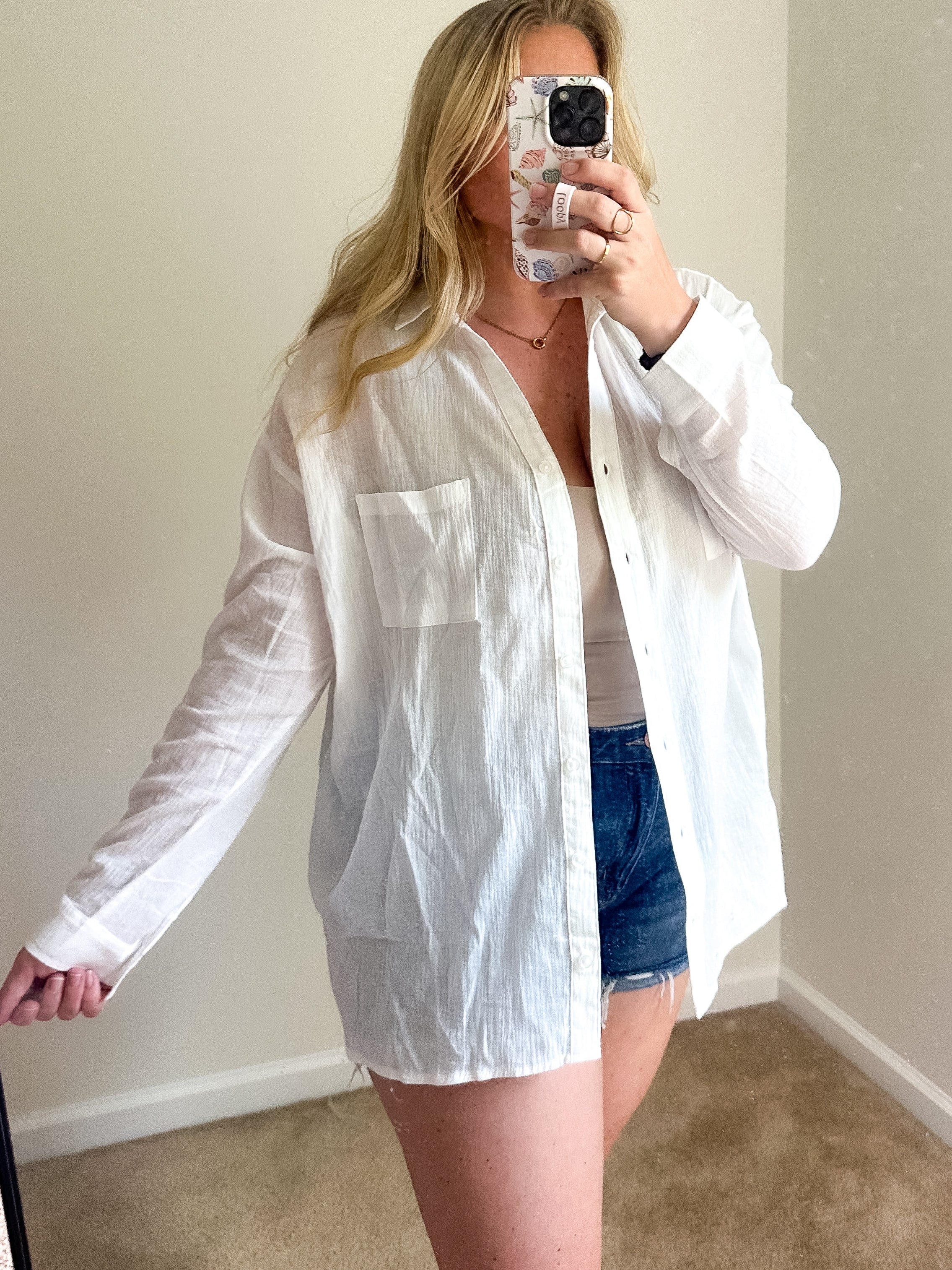 A woman takes a mirror selfie wearing an oversized white long-sleeve button-up shirt over a white top and denim shorts in a room with beige walls and carpet.