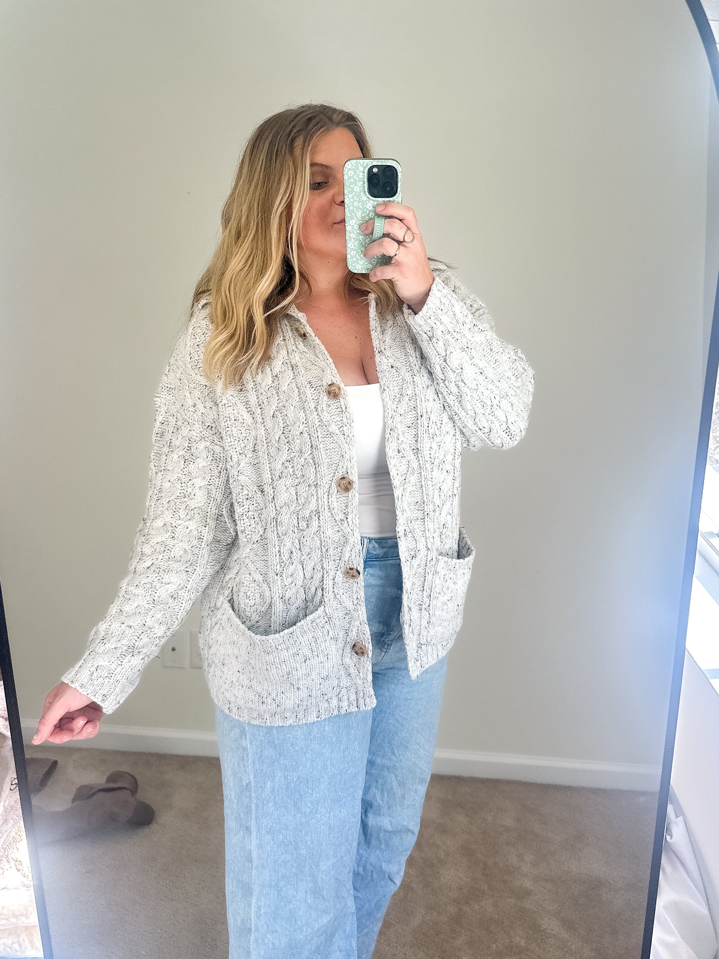 Woman taking a mirror selfie wearing a patterned cardigan and light blue jeans.