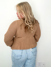 Person wearing a brown knitted sweater and light blue jeans against a white wall.