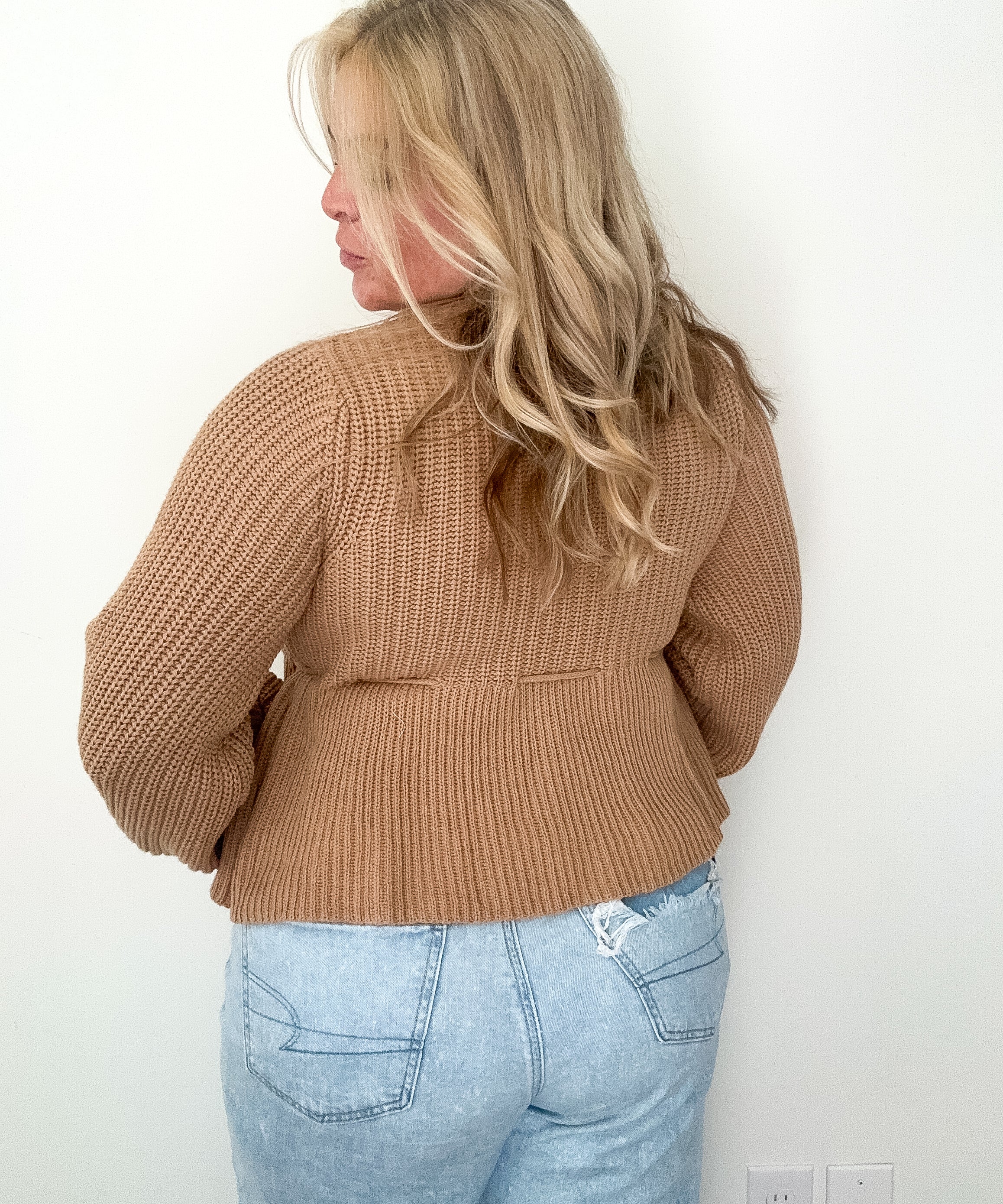 Person wearing a brown knitted sweater and light blue jeans against a white wall.