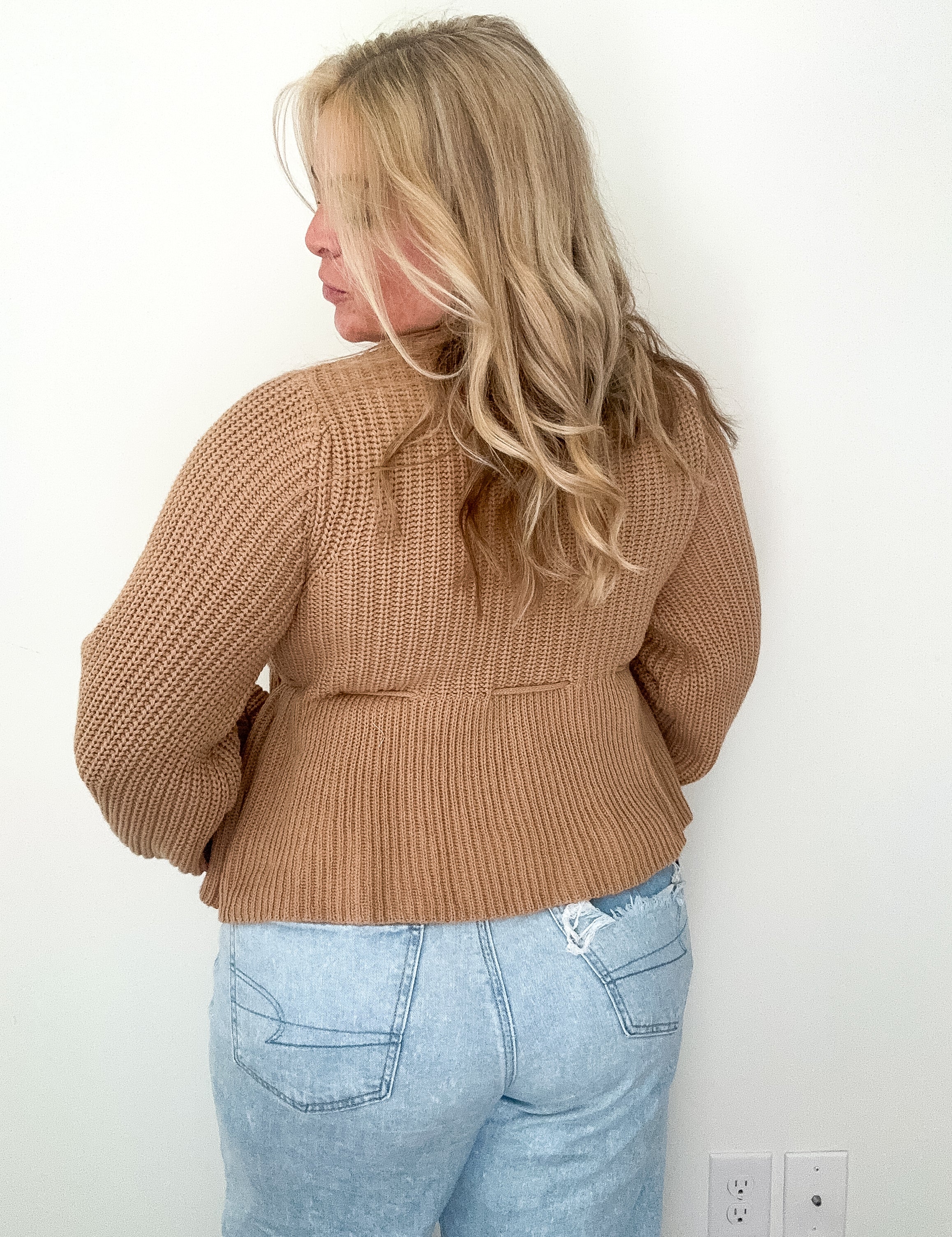 Person wearing a brown knitted sweater and light blue jeans against a white wall.
