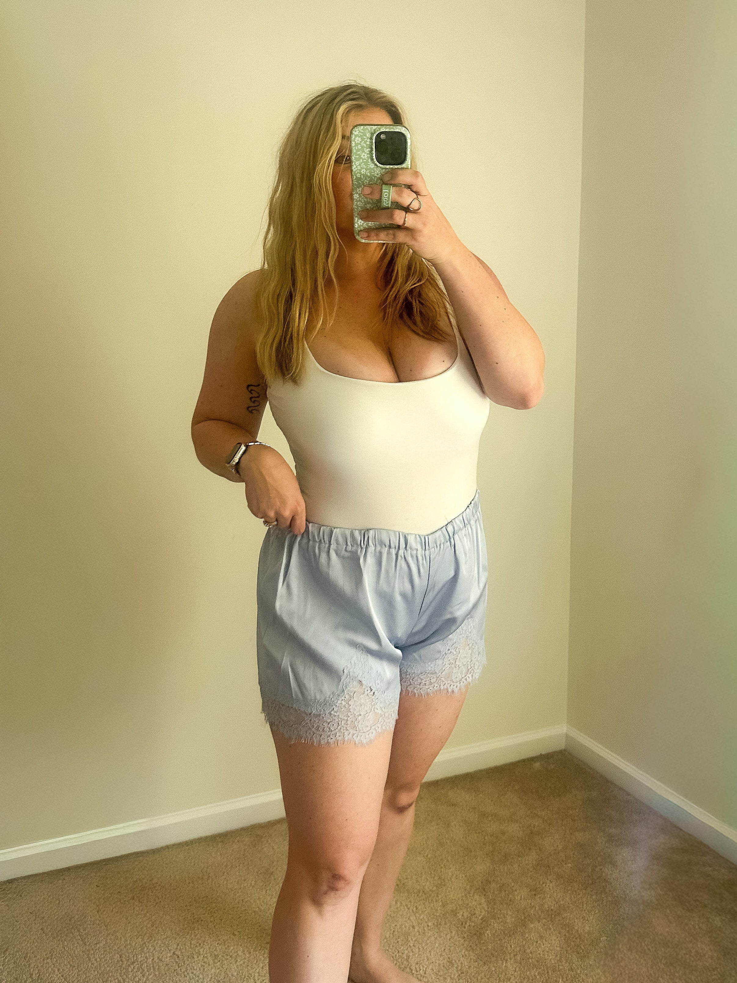 Person taking a mirror selfie wearing a white tank top and light blue shorts in a room with beige walls and carpet.