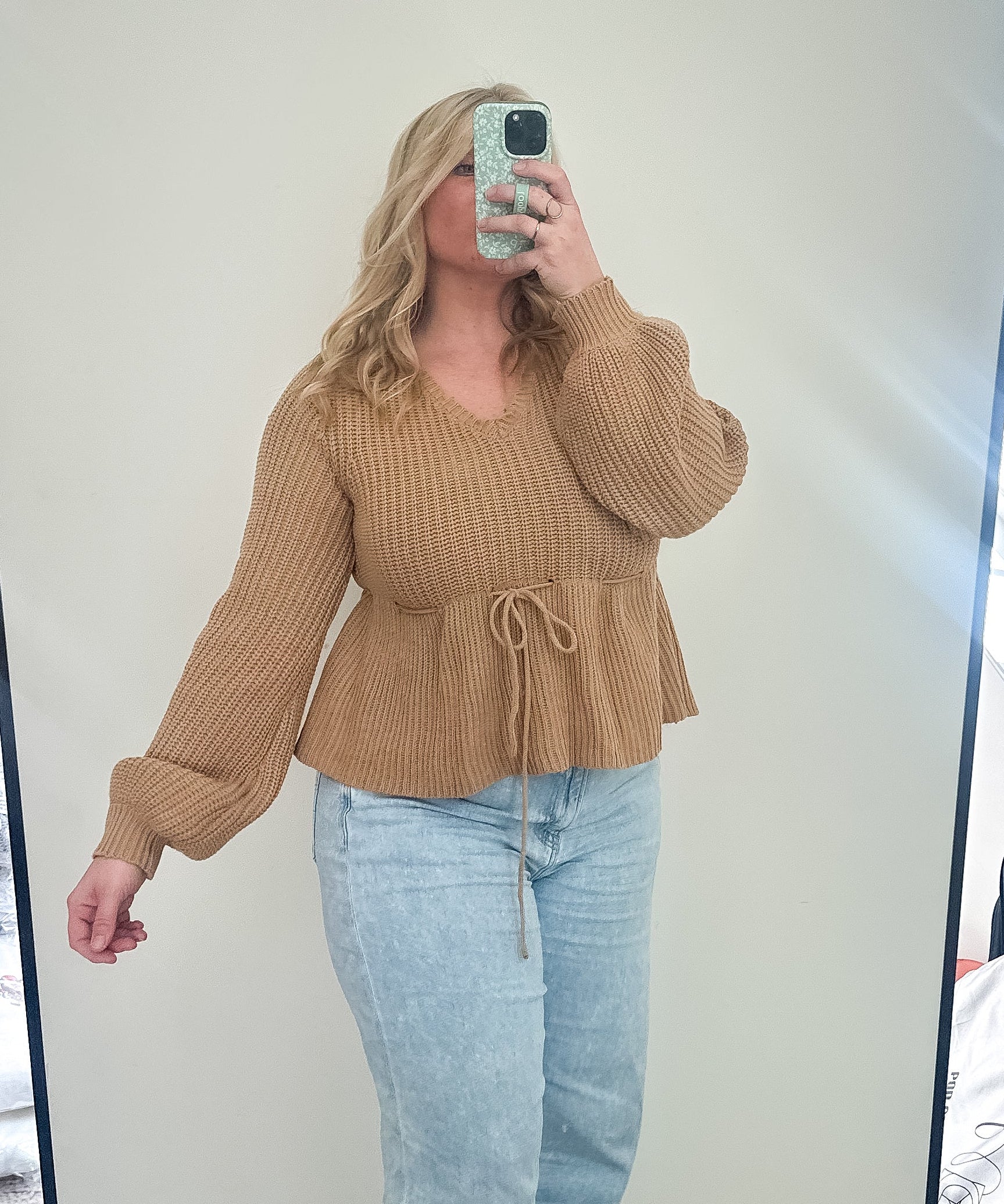 Person taking a mirror selfie wearing a brown sweater and light blue jeans in a room.