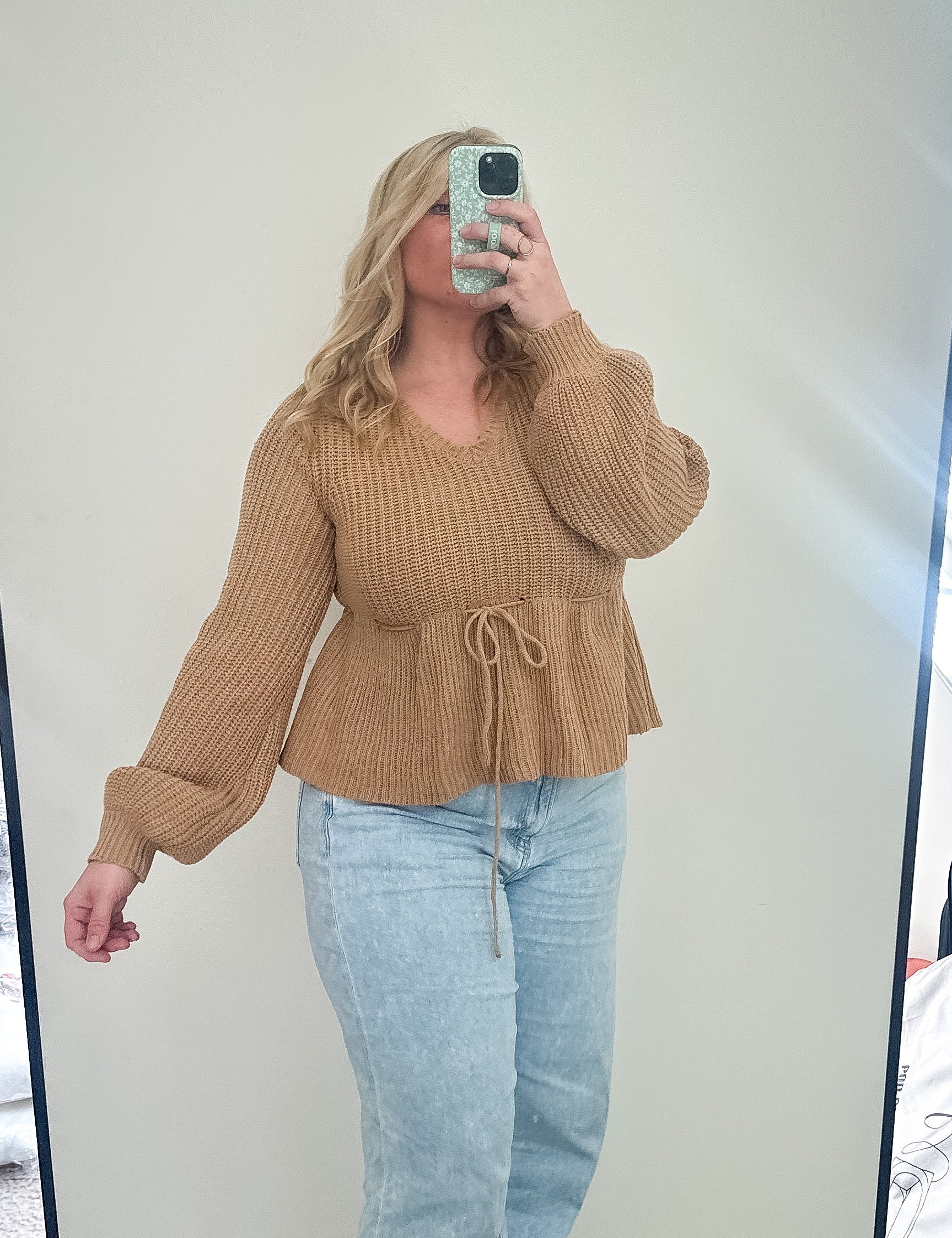 Person taking a mirror selfie wearing a brown sweater and light blue jeans in a room.