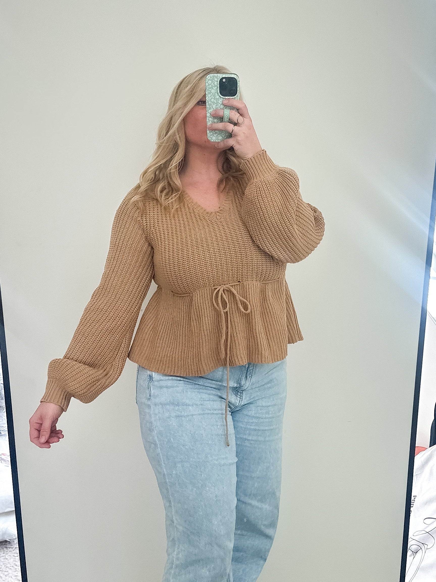 Person taking a mirror selfie wearing a brown sweater and light blue jeans in a room.