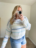 A woman takes a mirror selfie wearing a white knit sweater with horizontal stripes in light blue, yellow, and green, paired with denim shorts in a neutral indoor room.