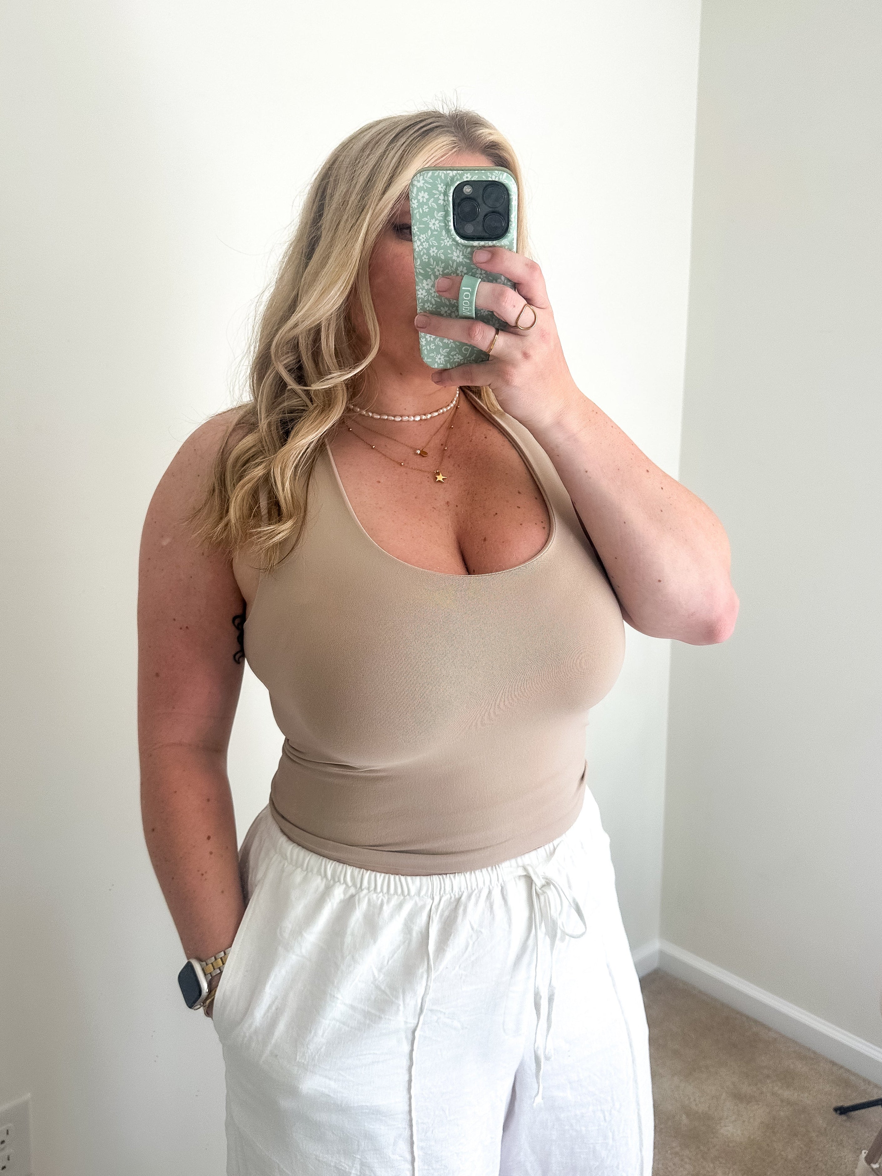 Woman taking a mirror selfie wearing a beige tank top and white pants.