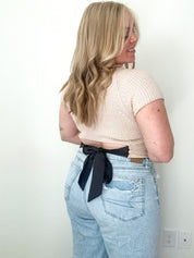 Person wearing a beige ribbed top and light blue jeans with a black ribbon, standing against a white wall.