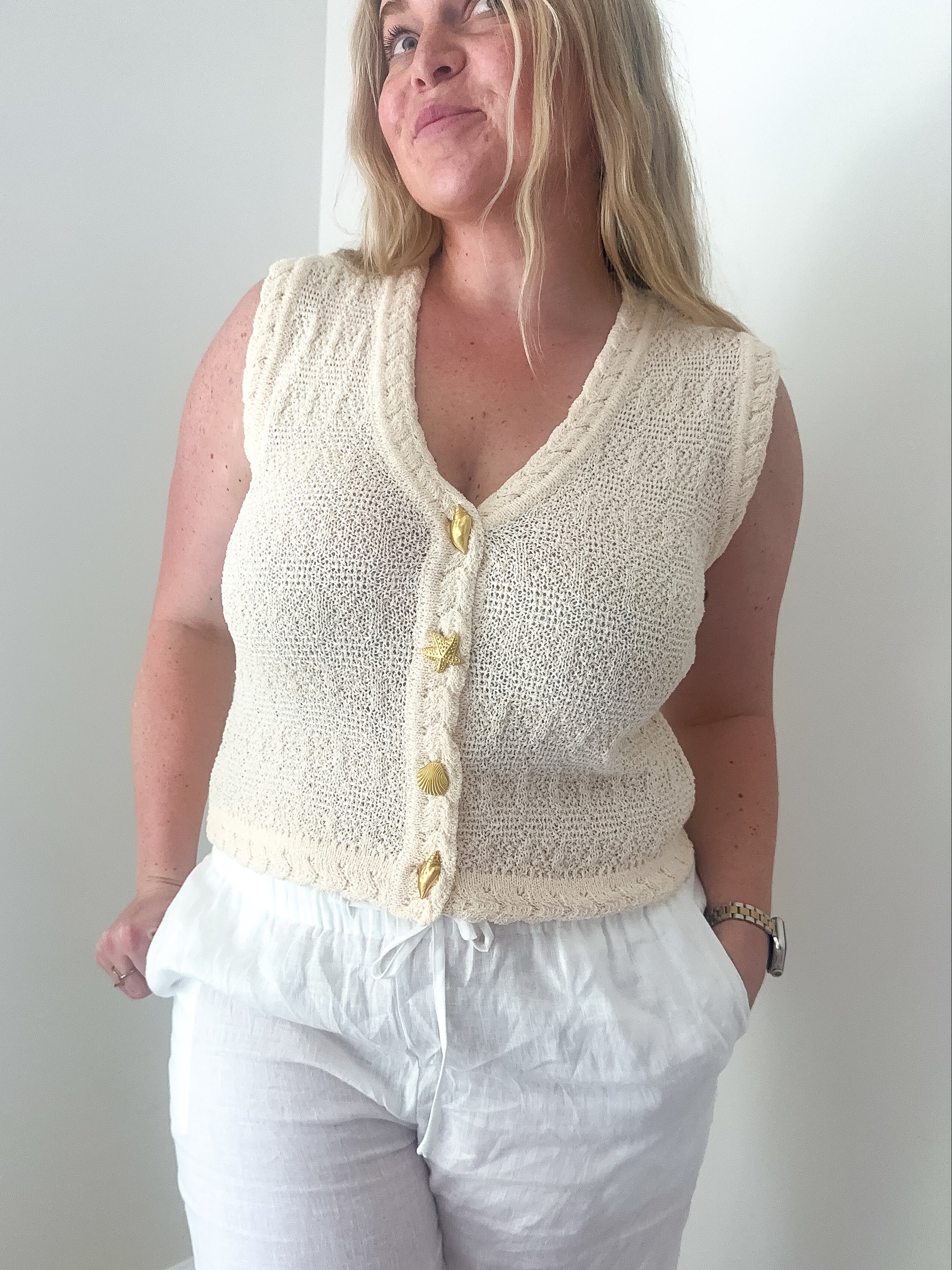 Woman wearing a cream knitted vest over a white top and pants against a plain background