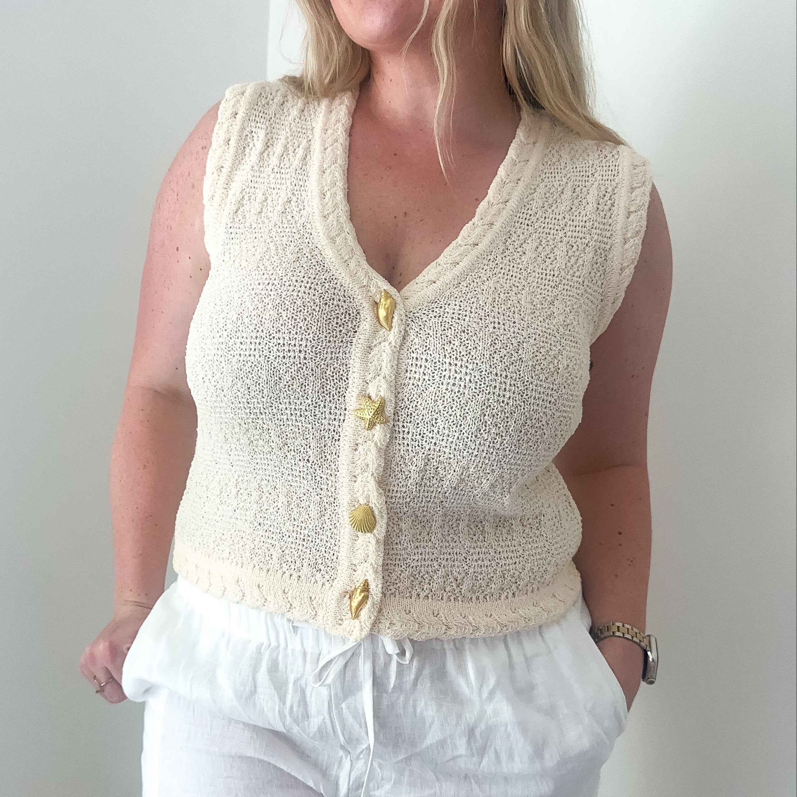 Woman wearing a cream knitted vest over a white top and pants against a plain background