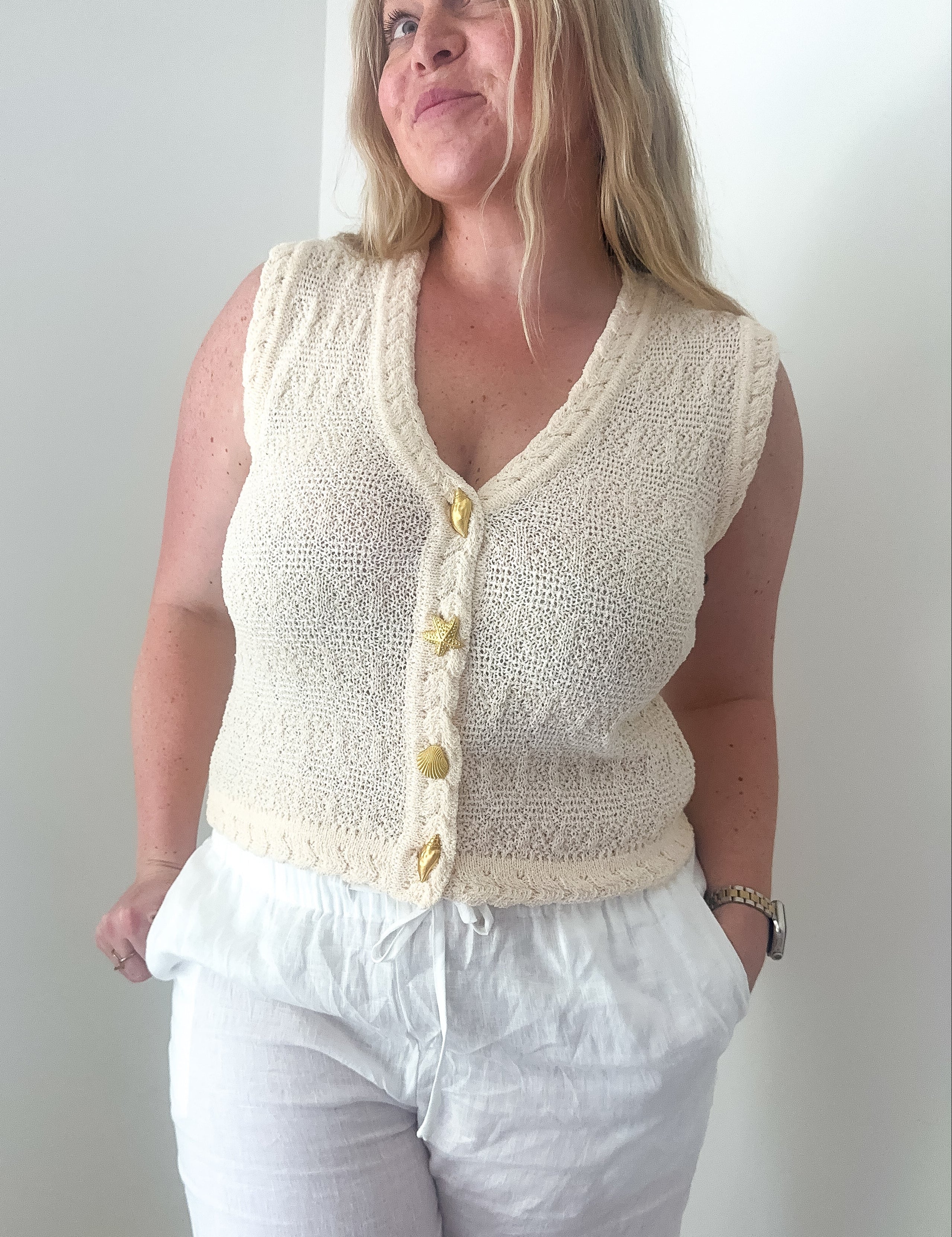 Woman wearing a cream knitted vest over a white top and pants against a plain background