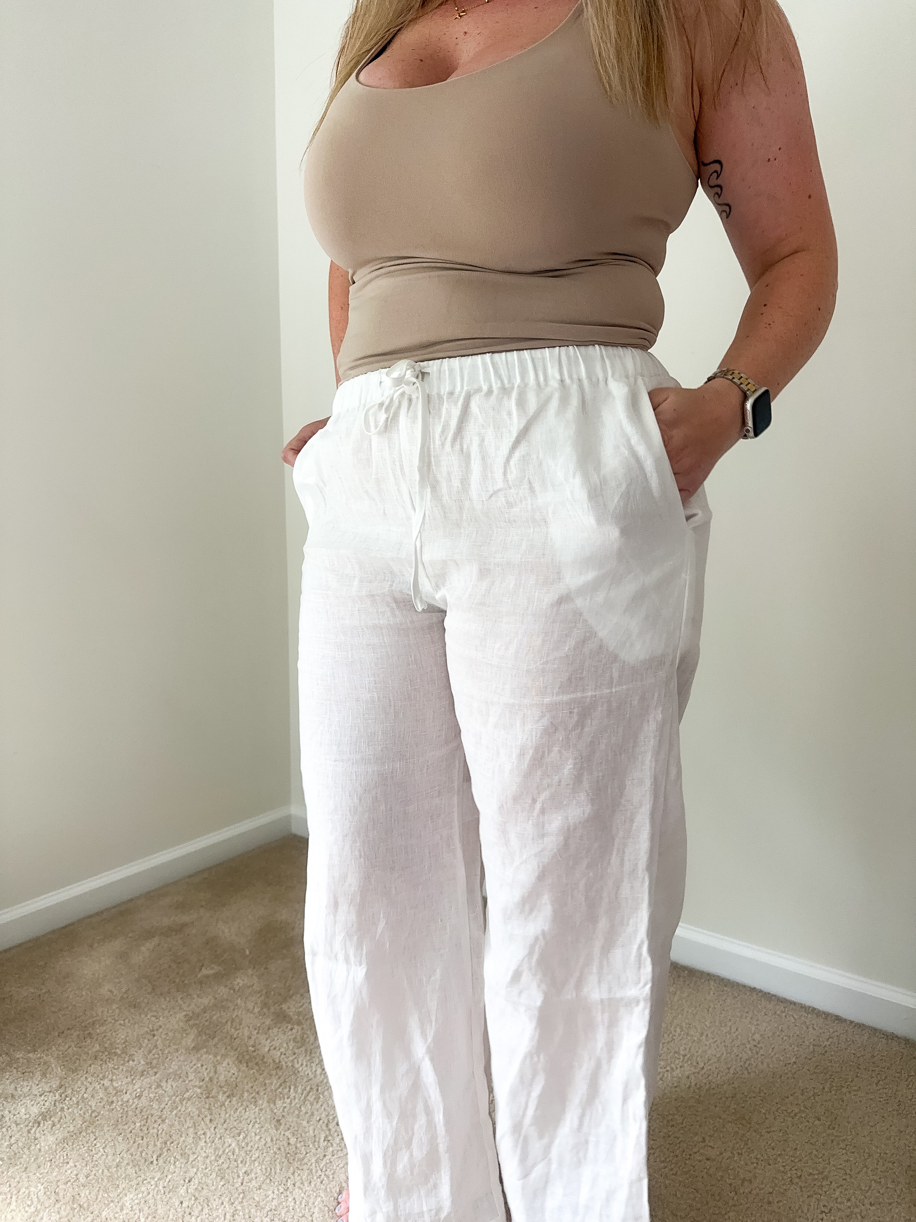 Person wearing a beige top and white pants against a plain wall.