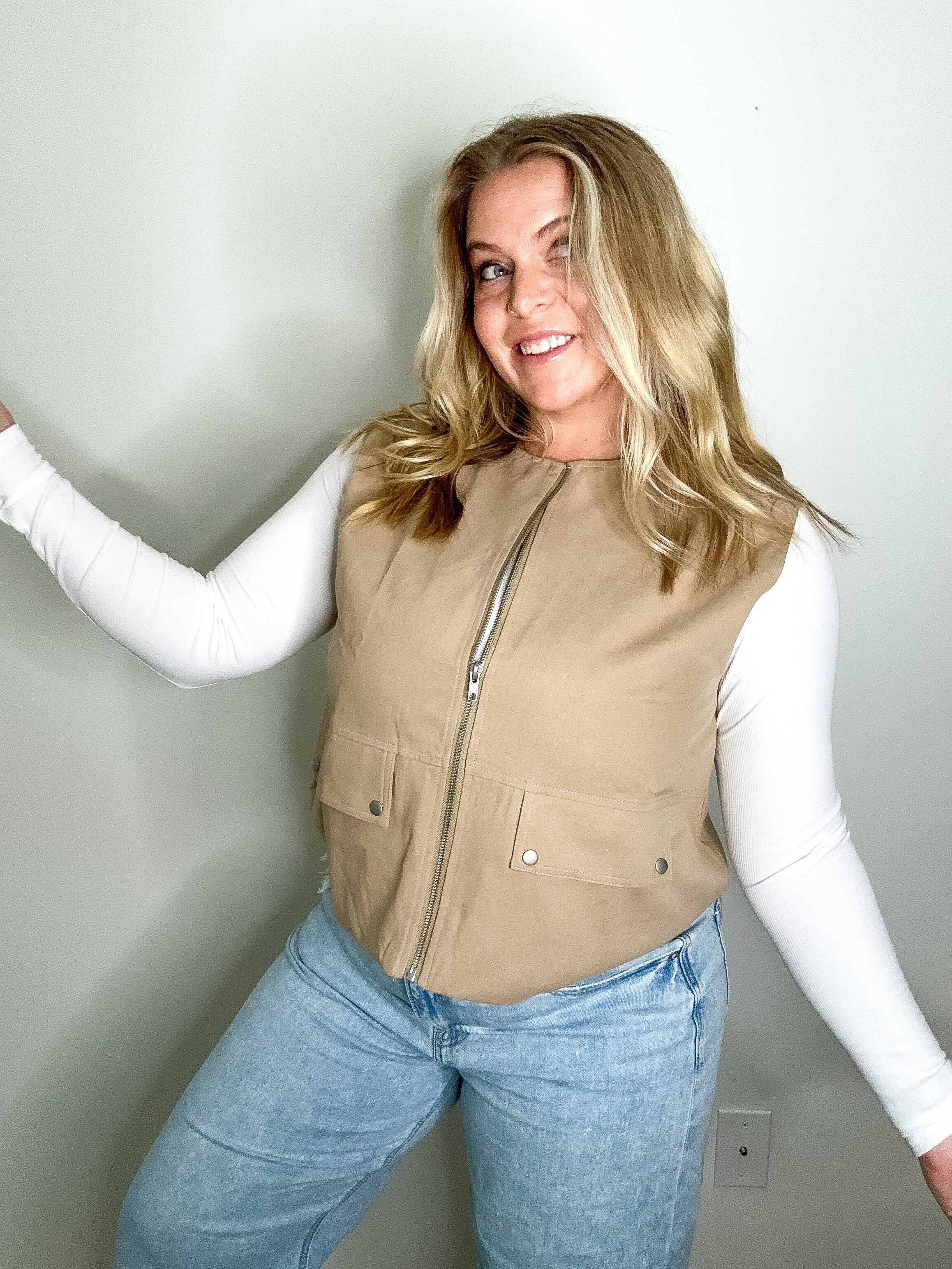 Woman wearing a beige vest over a white long-sleeve shirt and blue jeans against a plain background
