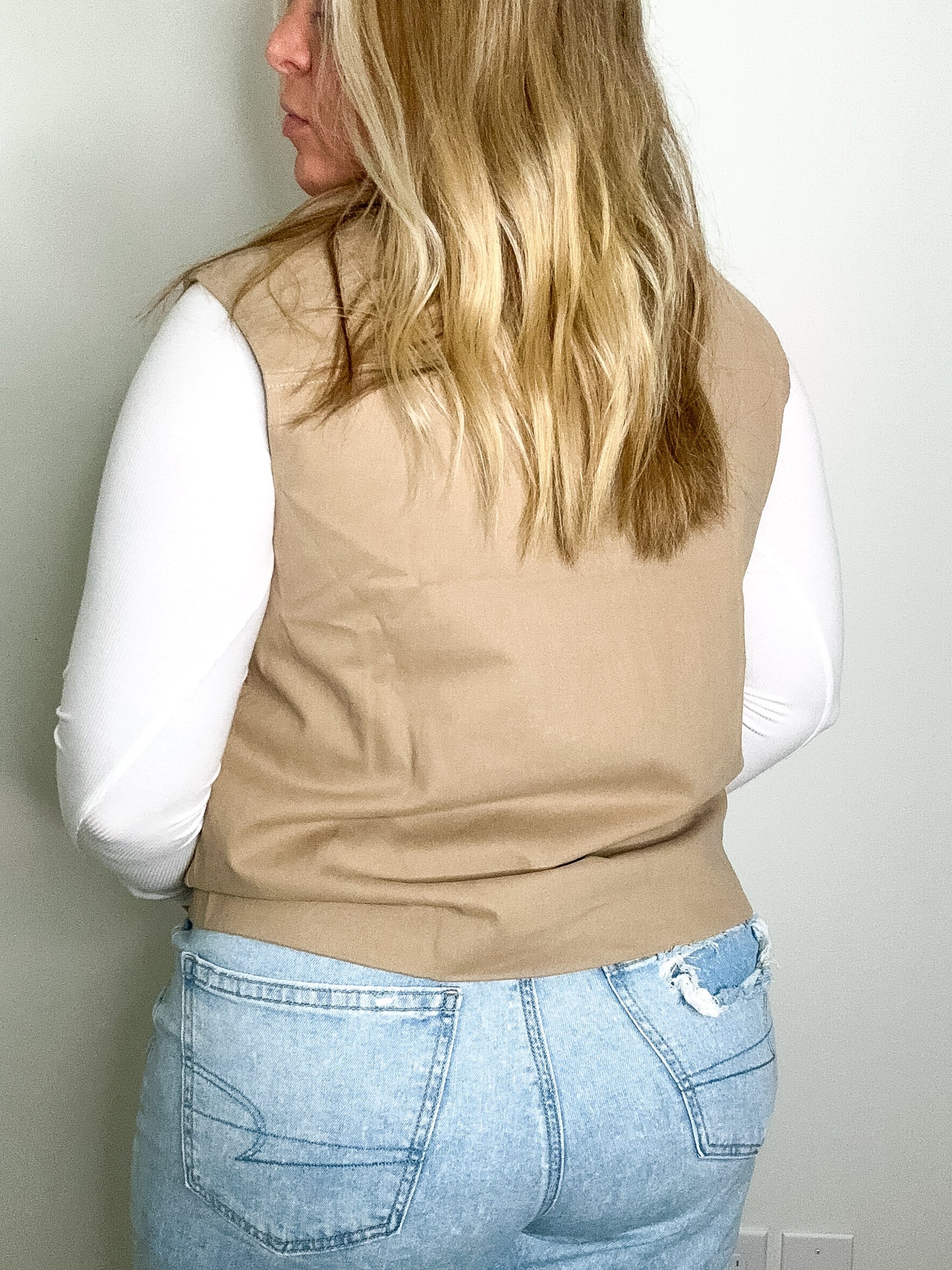 Person wearing a beige vest over a white shirt and blue jeans against a plain wall.