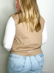 Person wearing a beige vest over a white shirt and blue jeans against a plain wall.