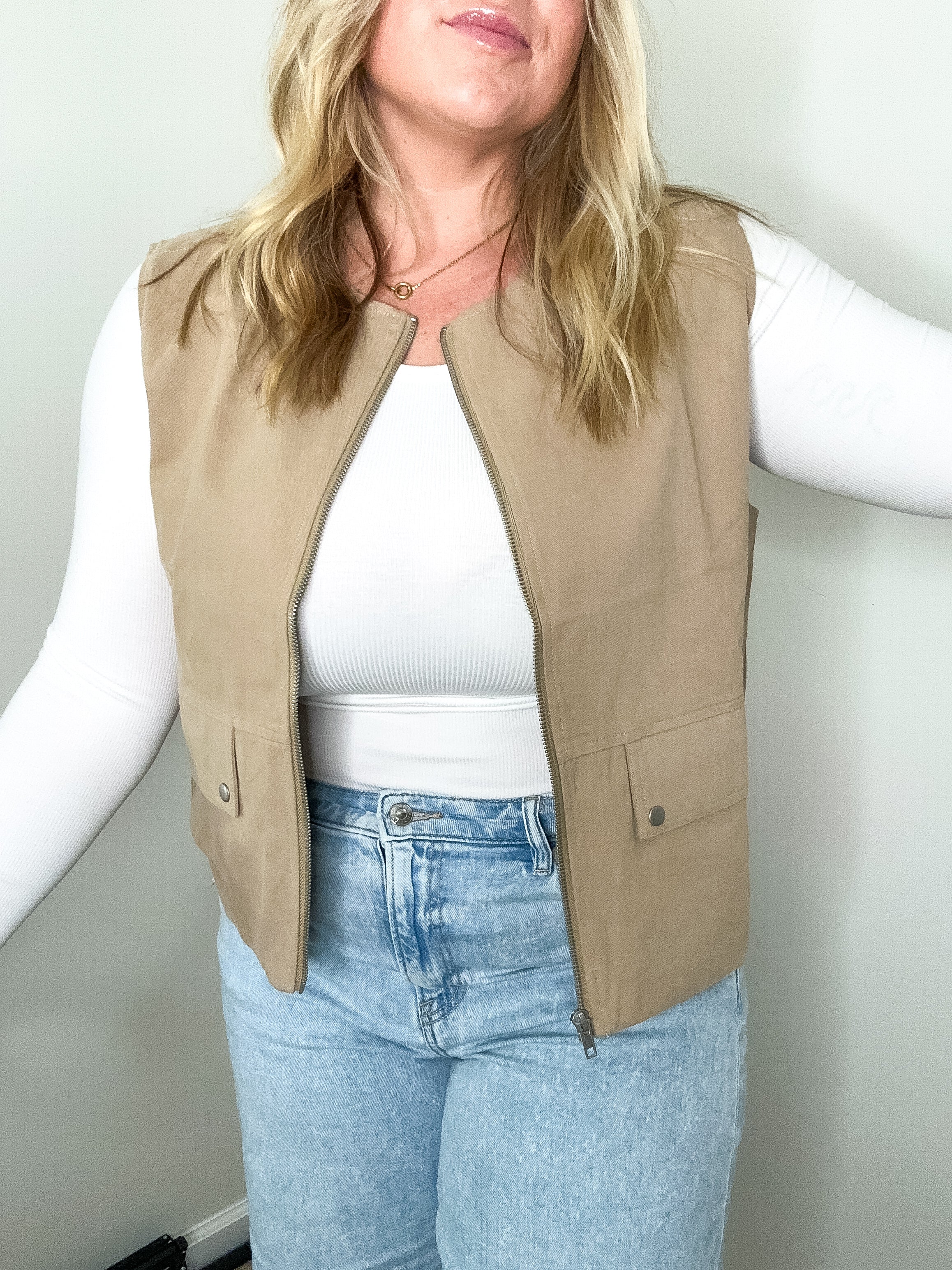 Person wearing a beige vest over a white shirt and blue jeans against a plain background