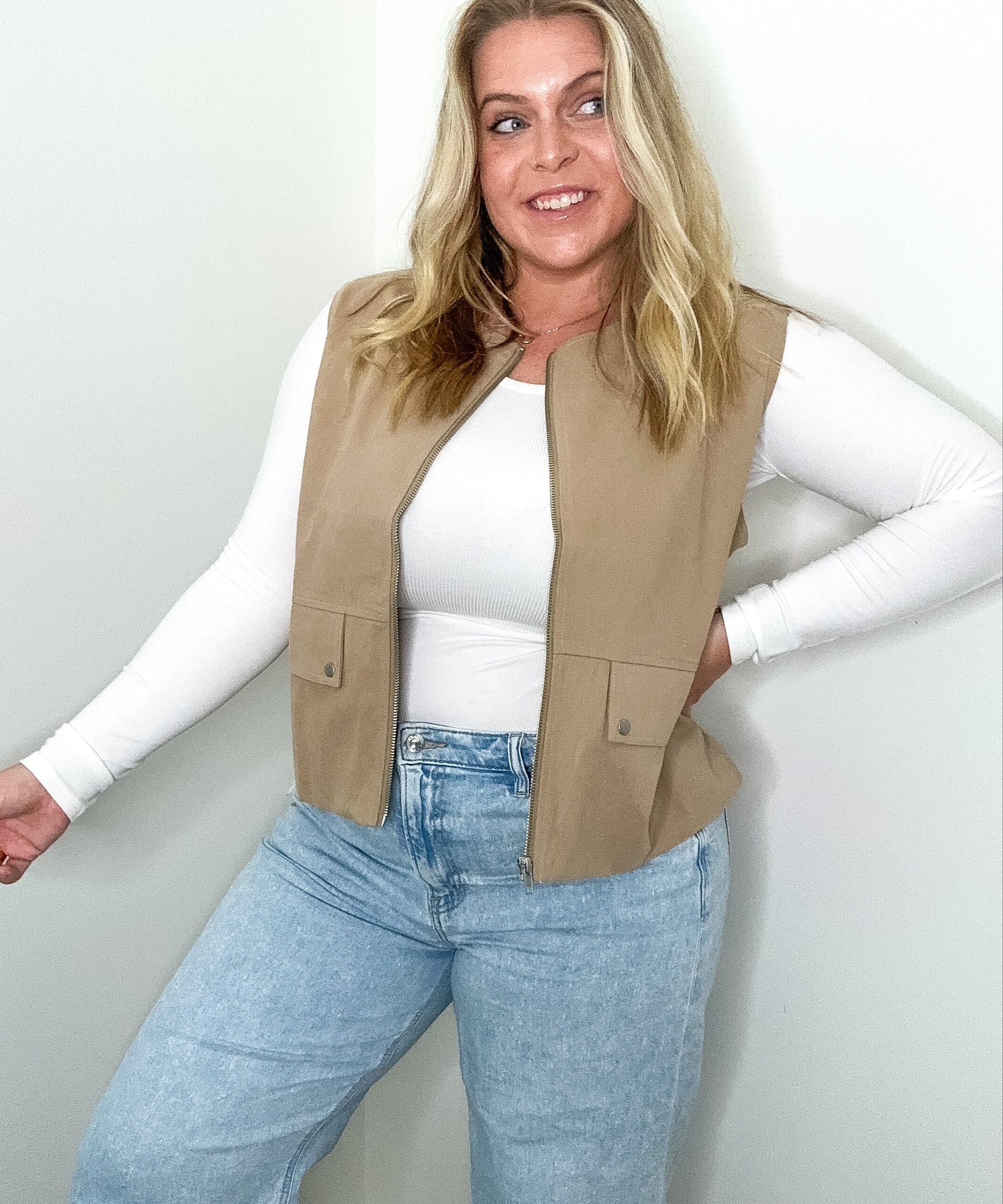 Woman wearing a beige vest, white long-sleeve shirt, and blue jeans against a plain background