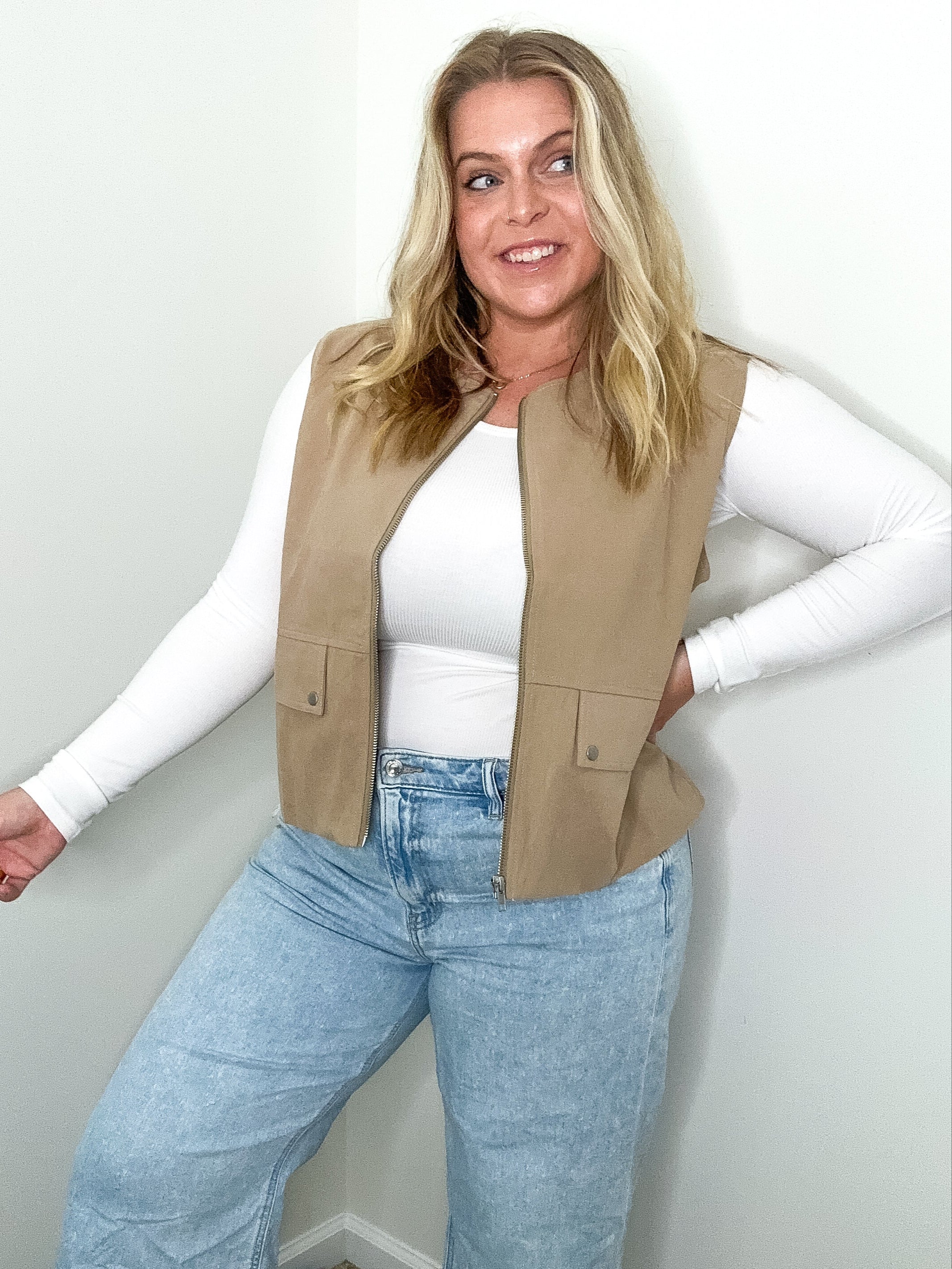 Woman wearing a beige vest, white long-sleeve shirt, and blue jeans against a plain background
