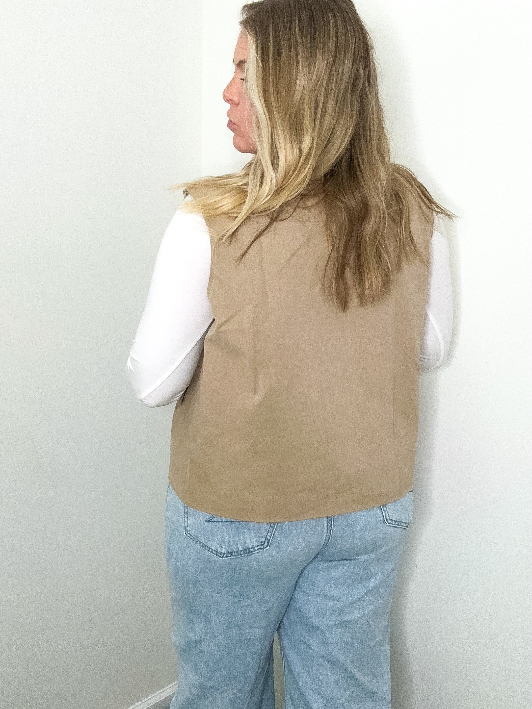 Person wearing a beige vest and light blue jeans against a white wall