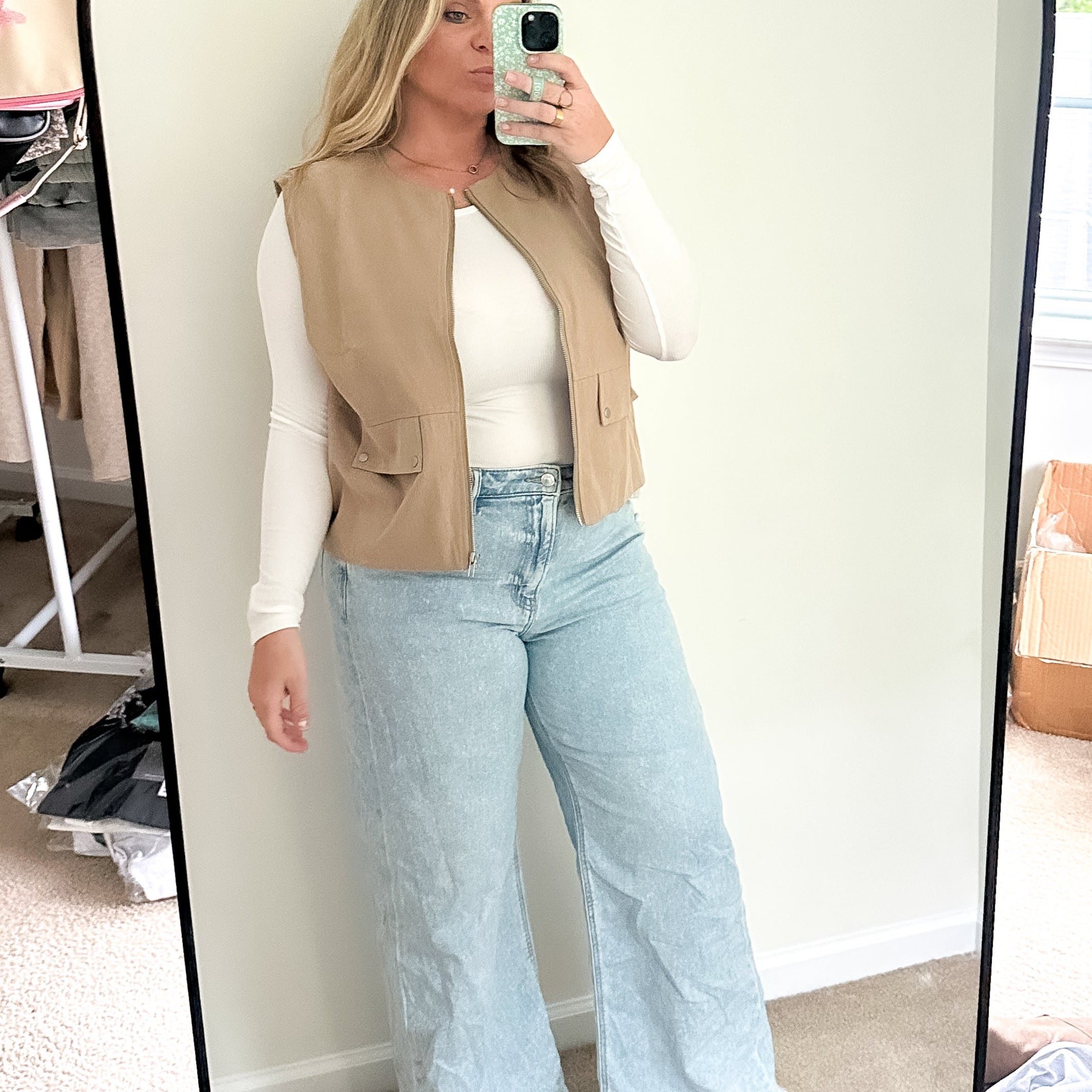 Person taking a mirror selfie wearing a beige vest, white shirt, and light blue jeans in a room with a window and boxes.