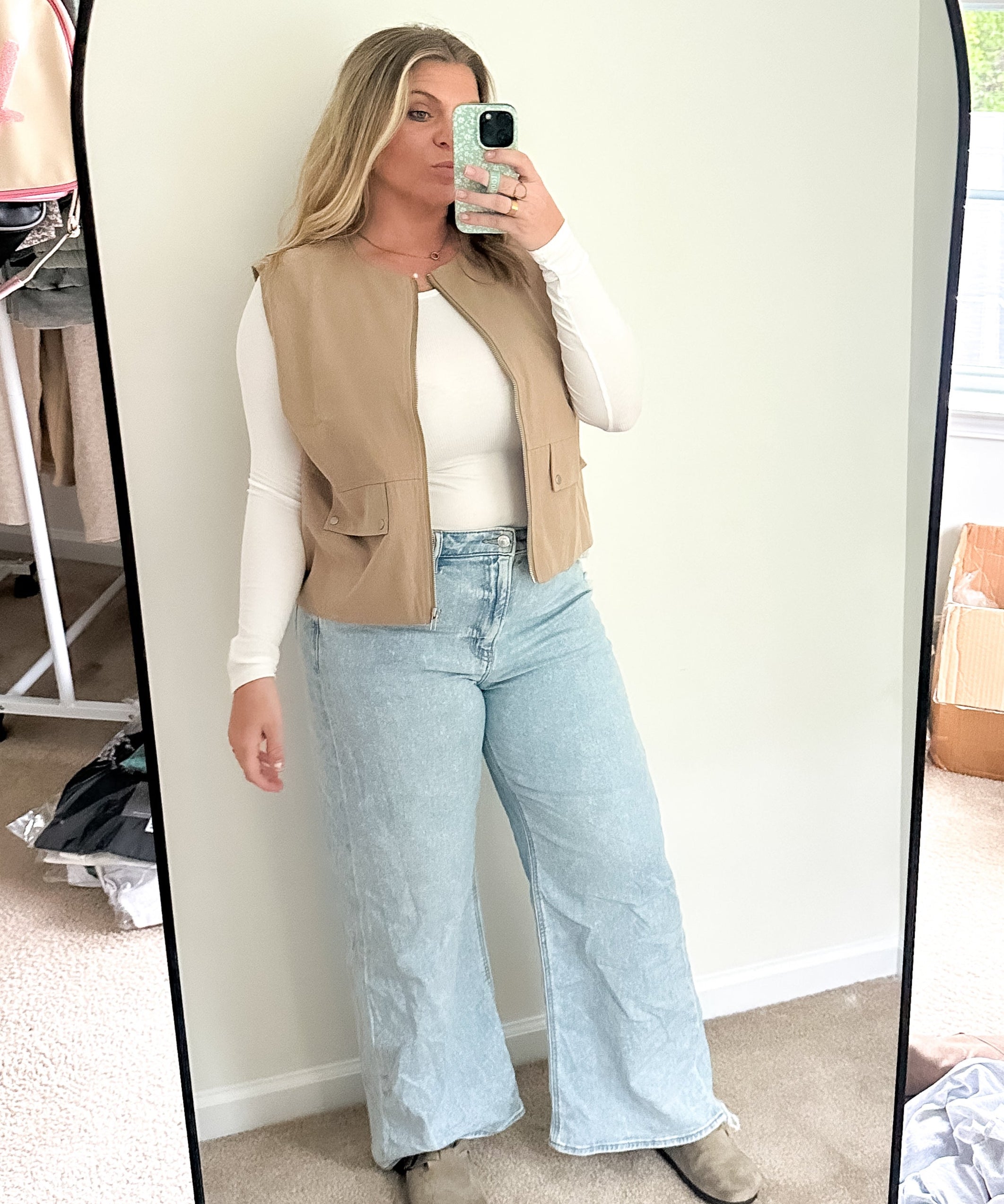 Person taking a mirror selfie wearing a beige vest, white shirt, and light blue jeans in a room with a window and boxes.