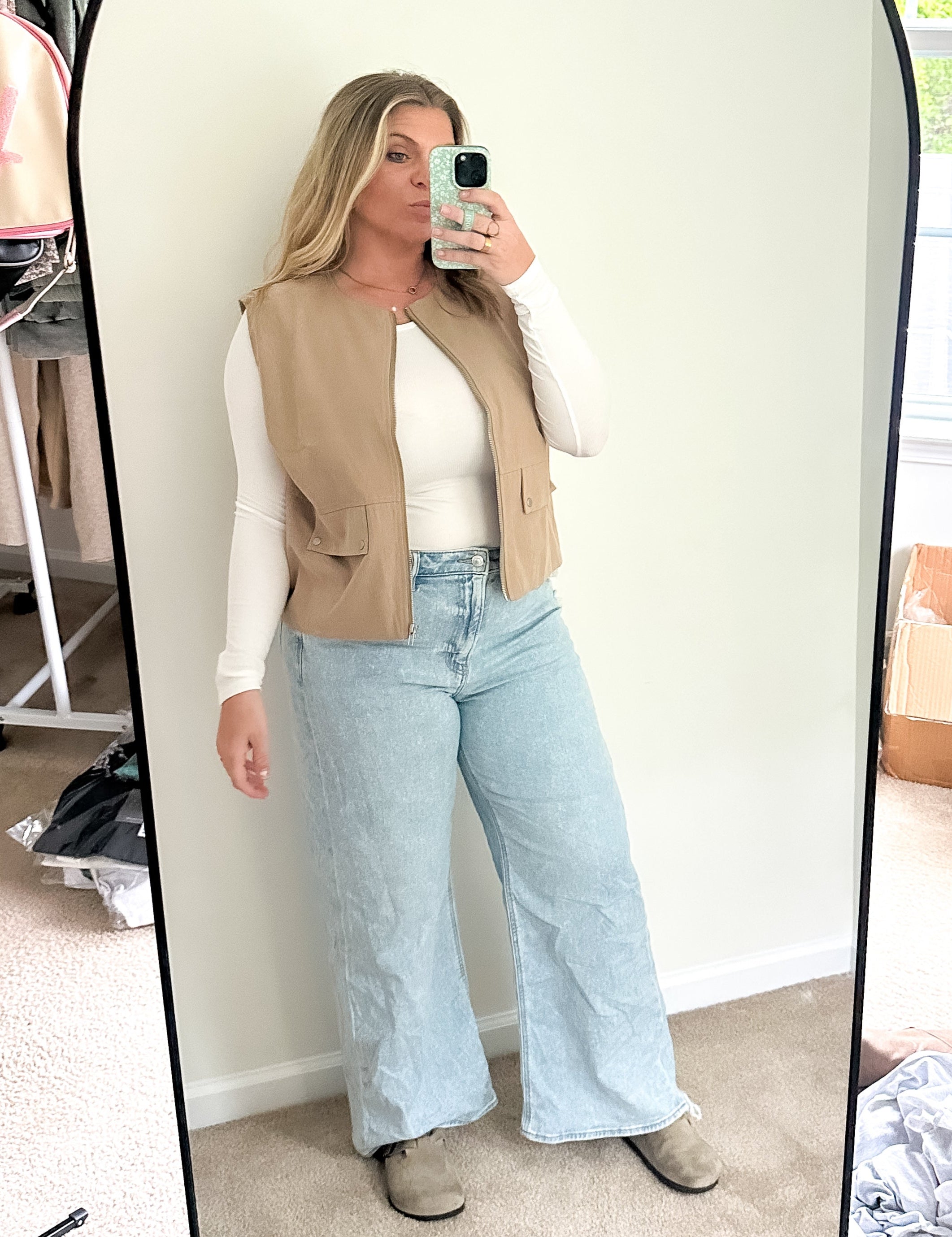 Person taking a mirror selfie wearing a beige vest, white shirt, and light blue jeans in a room with a window and boxes.